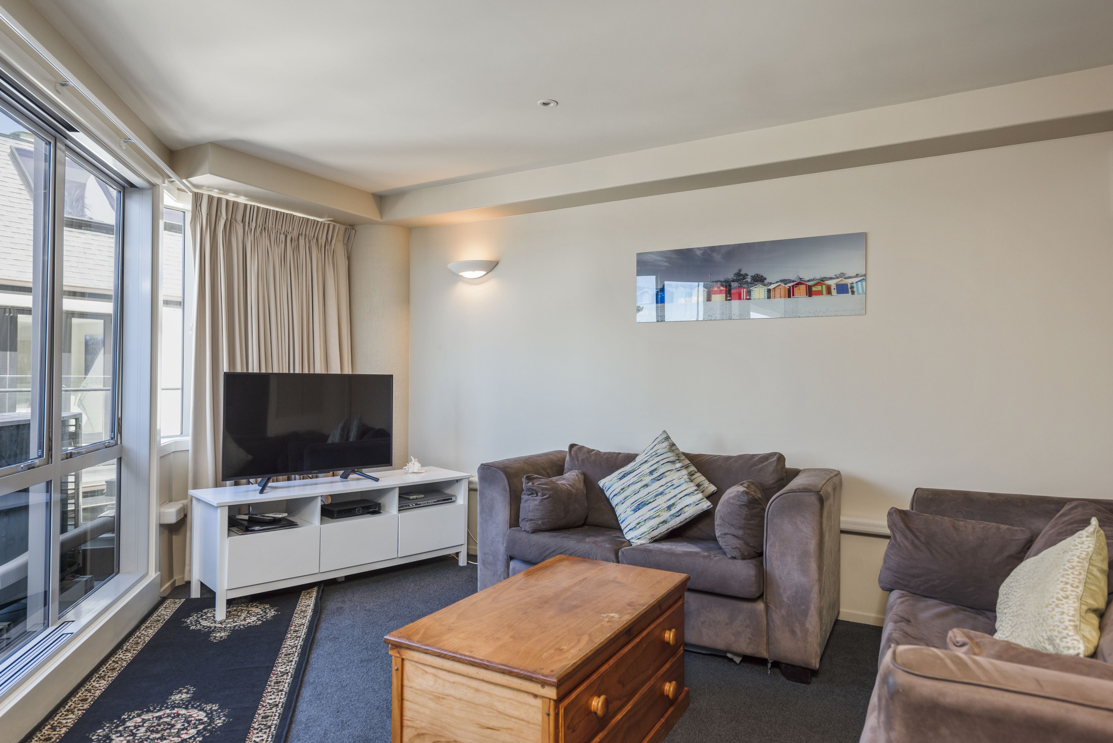 A living room with a flat-screen TV, two sofas, a wooden chest of drawers, and a large window.