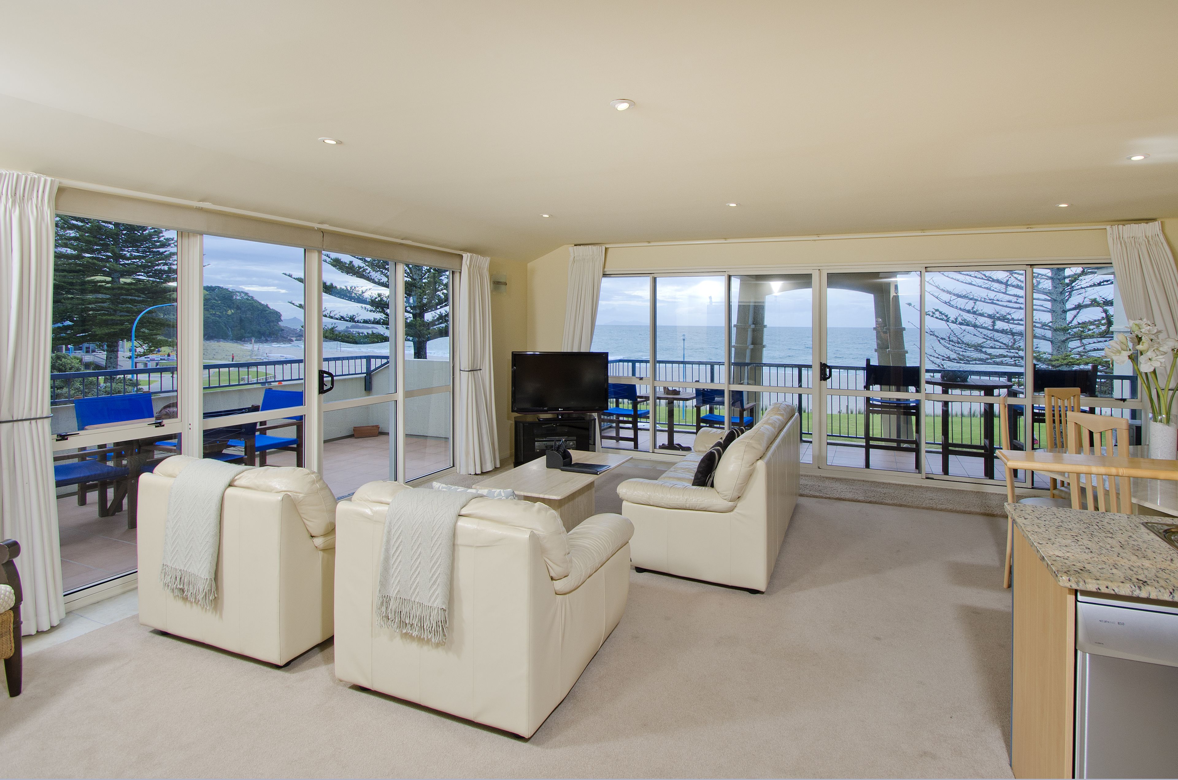 A spacious living area with two white sofas, a flat-screen TV, and a dining table with chairs. Large windows offer a view of the sea and a balcony with a railing.