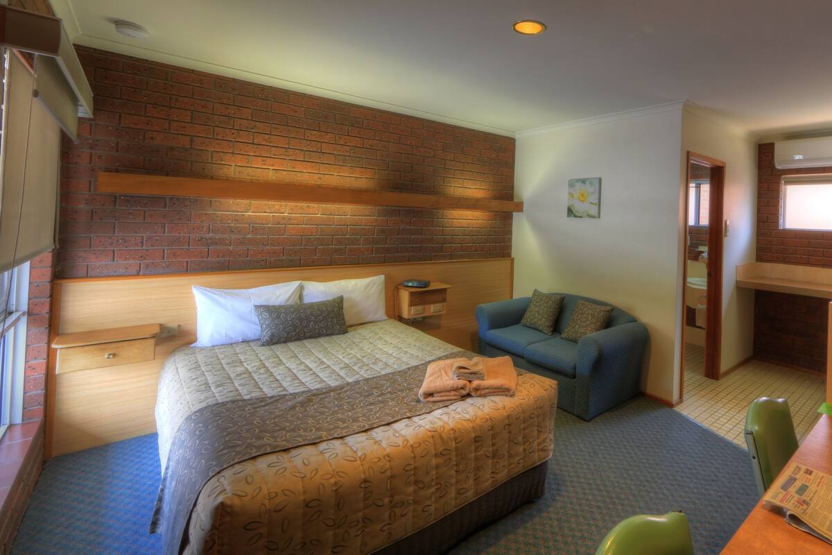A hotel room with a large bed, a blue sofa, a wooden desk with a television, and a brick wall.