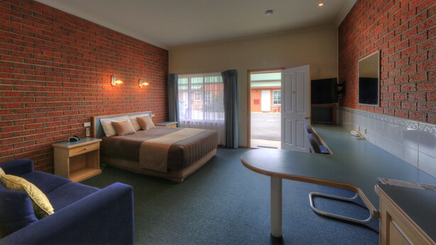 Quarto de hotel com parede de tijolos, cama com cabeceira, escrivaninha com cadeira, televisão e banheiro visível por uma porta aberta.