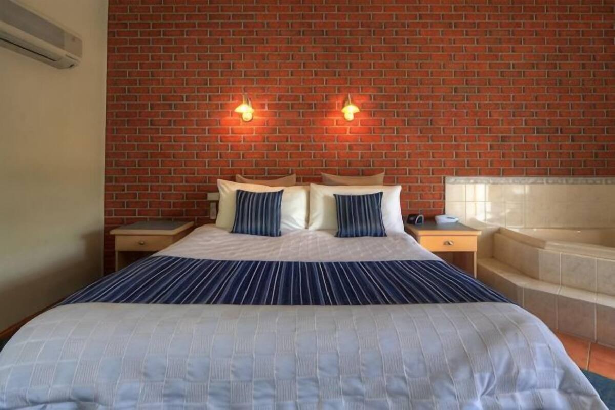 A hotel room with a brick wall, a bed with a striped bedspread, two bedside lamps, a nightstand, and a bathtub.