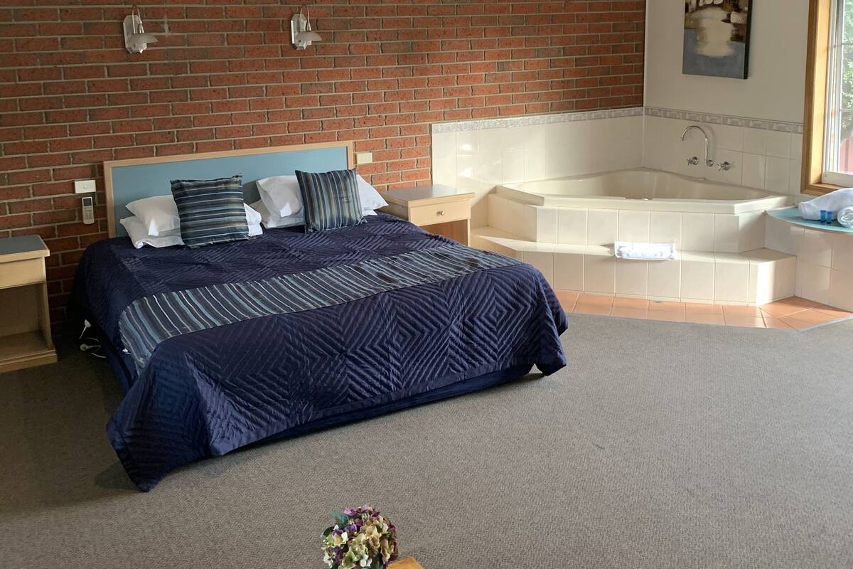 A modern bedroom with a brick wall, a large bed with a blue bedspread, a wooden coffee table with a plant, and a white bathtub in the corner.