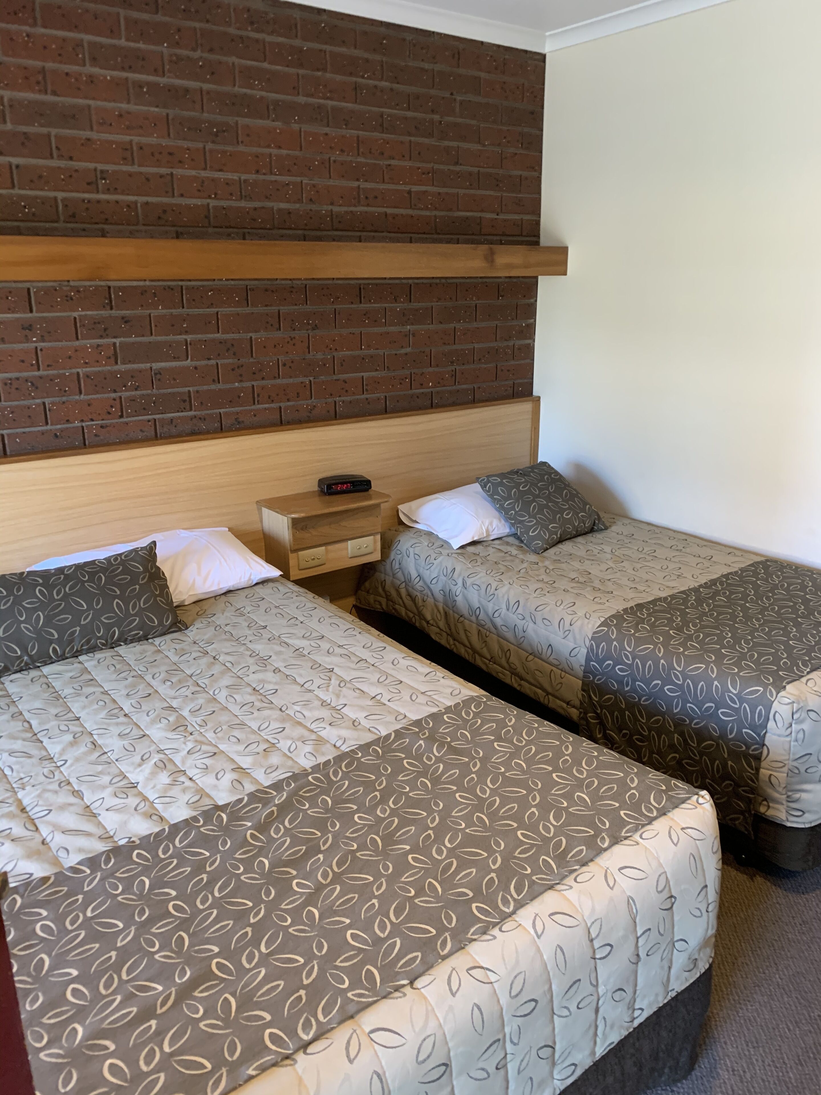 A hotel room with two beds, a brick-patterned headboard, a wooden nightstand, and a digital clock.