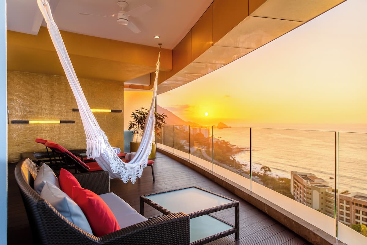 A balcony with a hammock, wicker furniture, and a view of the sunset over the ocean.