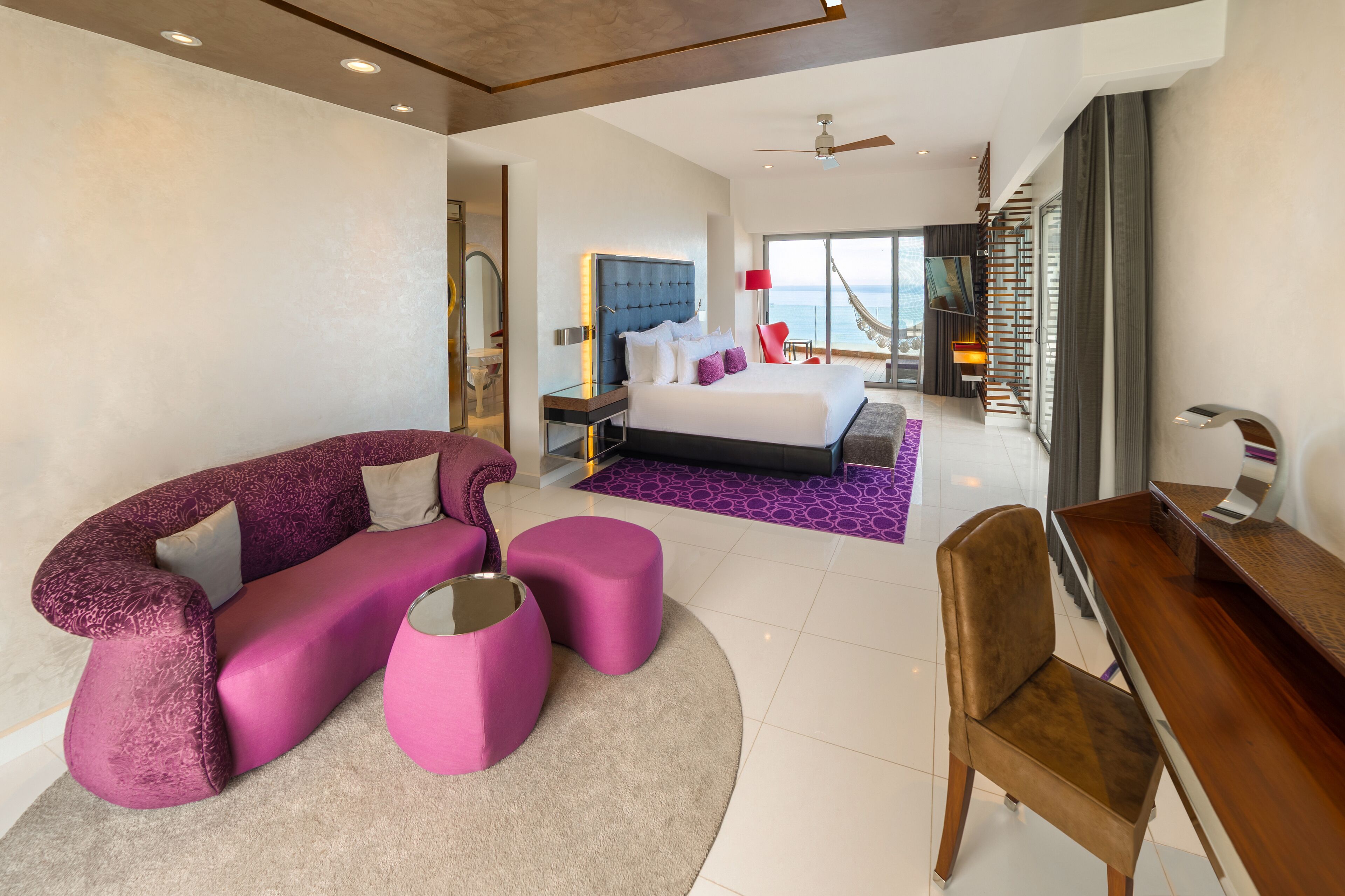 A modern hotel room with a curved purple sofa, a wooden desk, and a large bed with a blue headboard.