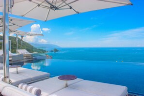 Una piscina infinita con sillones blancos y sombrillas, con vista al mar azul cristalino y montañas lejanas.