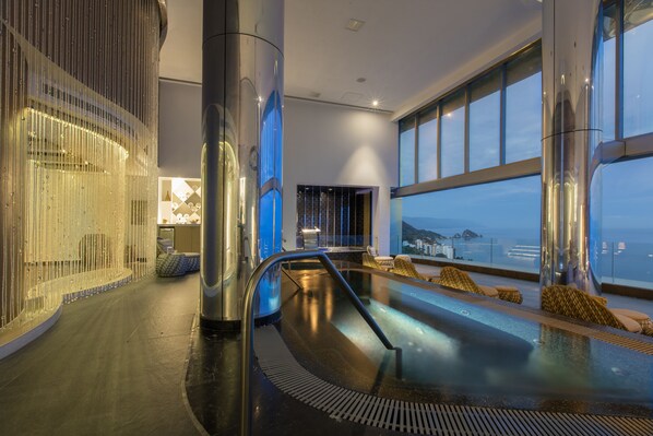 Un área de piscina interior moderna con barrera de cristal, sillas de descanso y vista al océano.