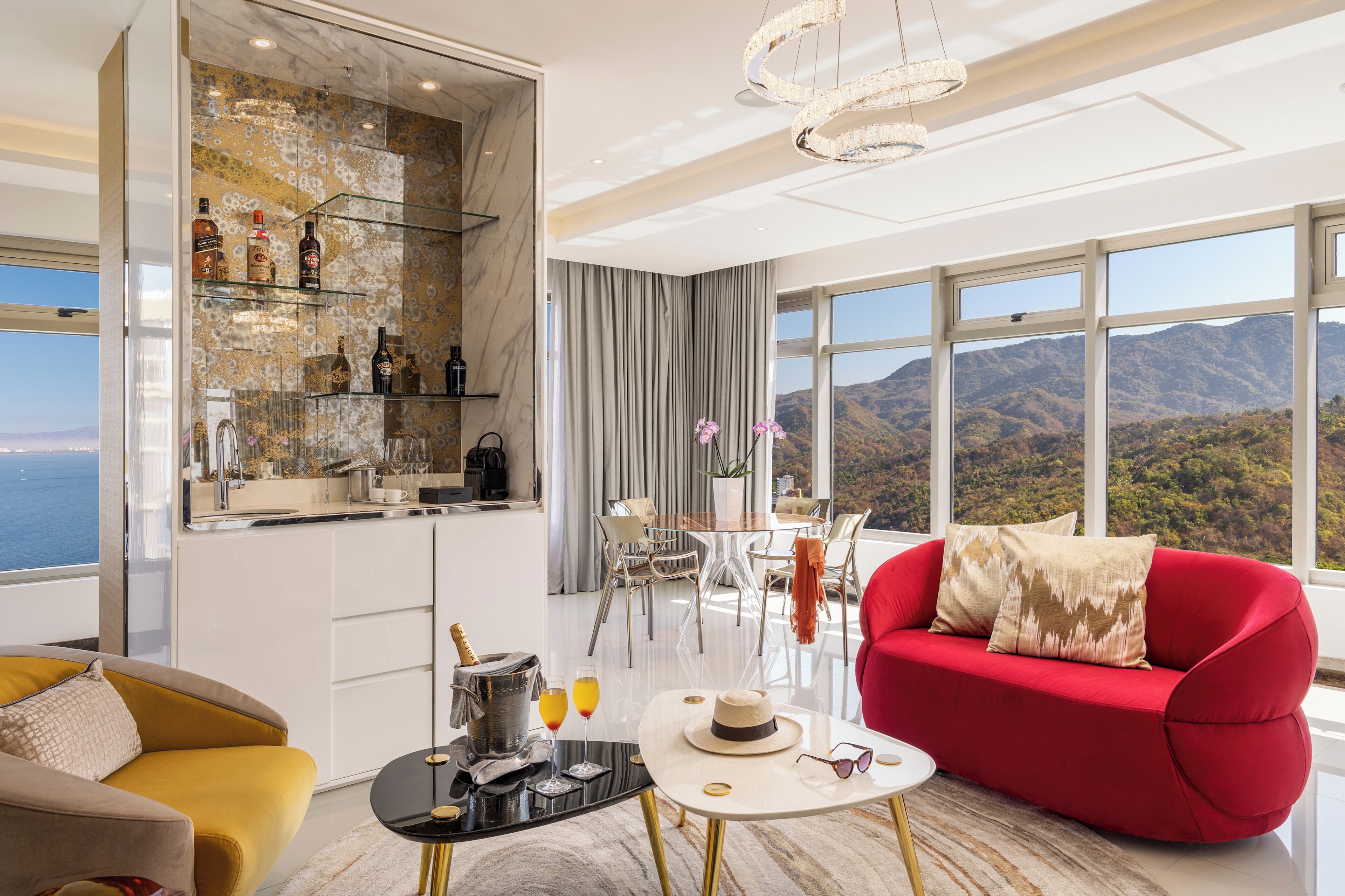 Una sala de estar moderna con un sofá rojo, un sillón amarillo, una barra de vidrio con botellas, un comedor y vistas a la montaña.