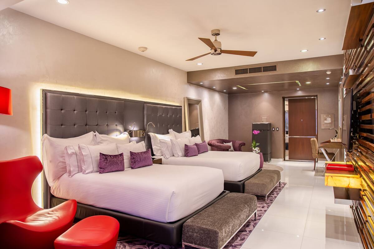 A hotel room with two beds, a red chair, a bench, a ceiling fan, and a minibar.
