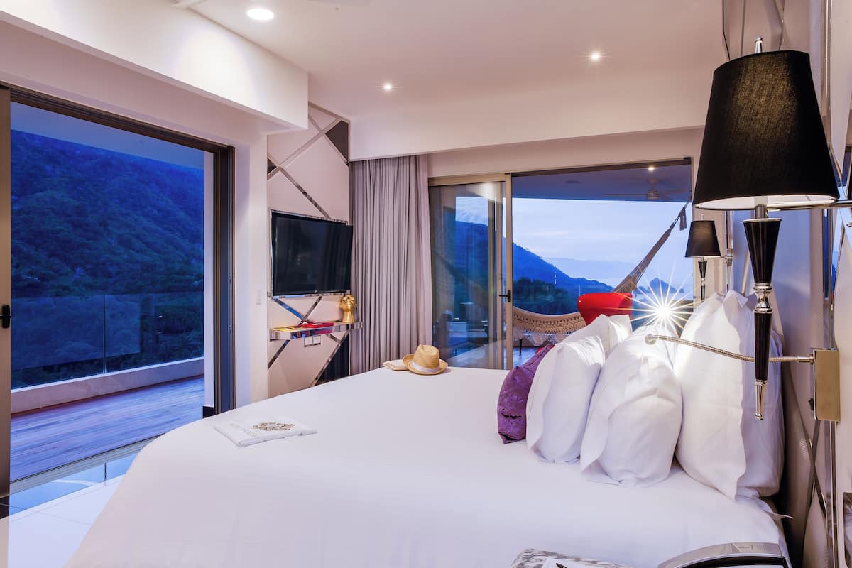 A modern hotel room with a large bed, a flat-screen TV, a desk, and a view of the mountains.
