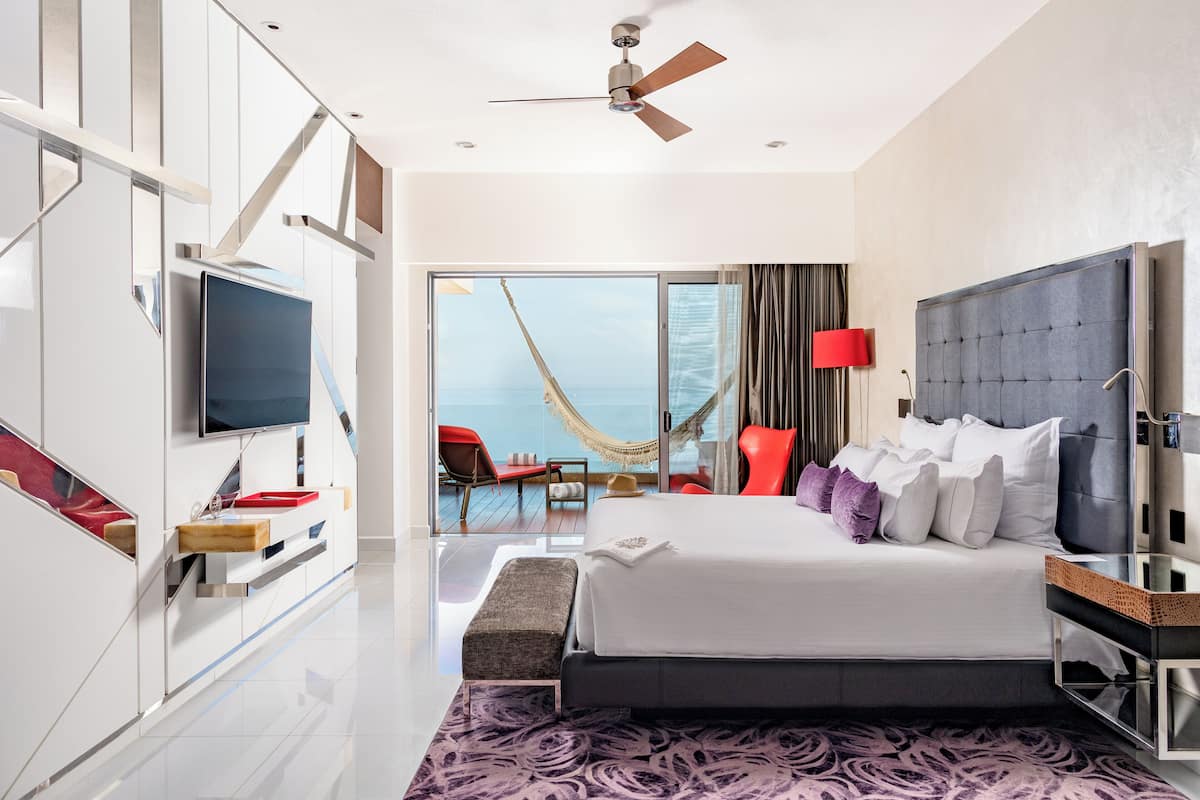 A modern hotel room with a large bed, a TV, a balcony with a view of the sea, and a ceiling fan.