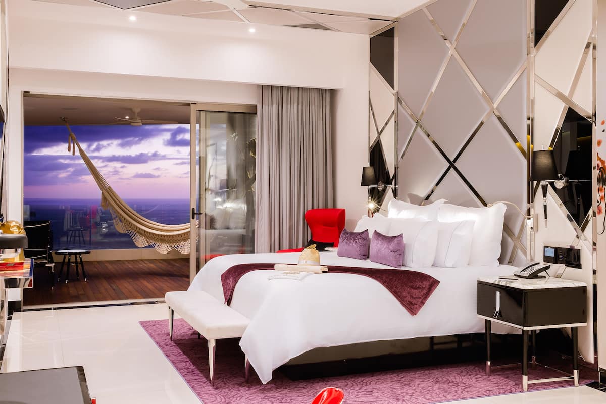A modern hotel room with a large bed, a red chair, a desk with a laptop, and a view of the city through a glass door.