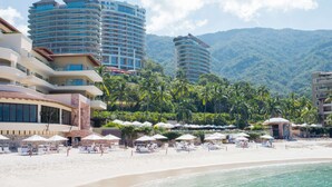 A beachfront hotel with multiple balconies, a sandy beach with umbrellas, and a mountainous backdrop. - Hotel Mousai Puerto Vallarta Adults Only (Puerto Vallarta)