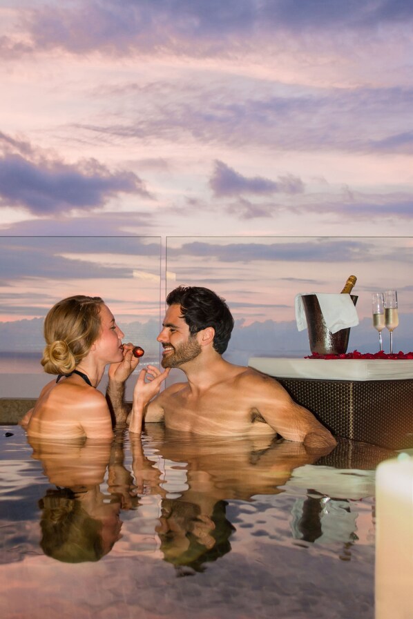 Una pareja en un jacuzzi con una botella de champán y copas, reflejándose en el agua con un atardecer de fondo.