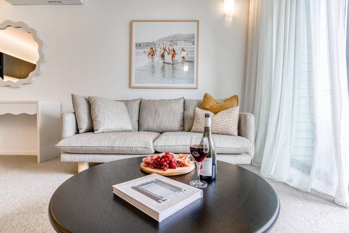 A modern living room with a sofa, a round coffee table, a book, a bottle of wine, and a plate of fruit.