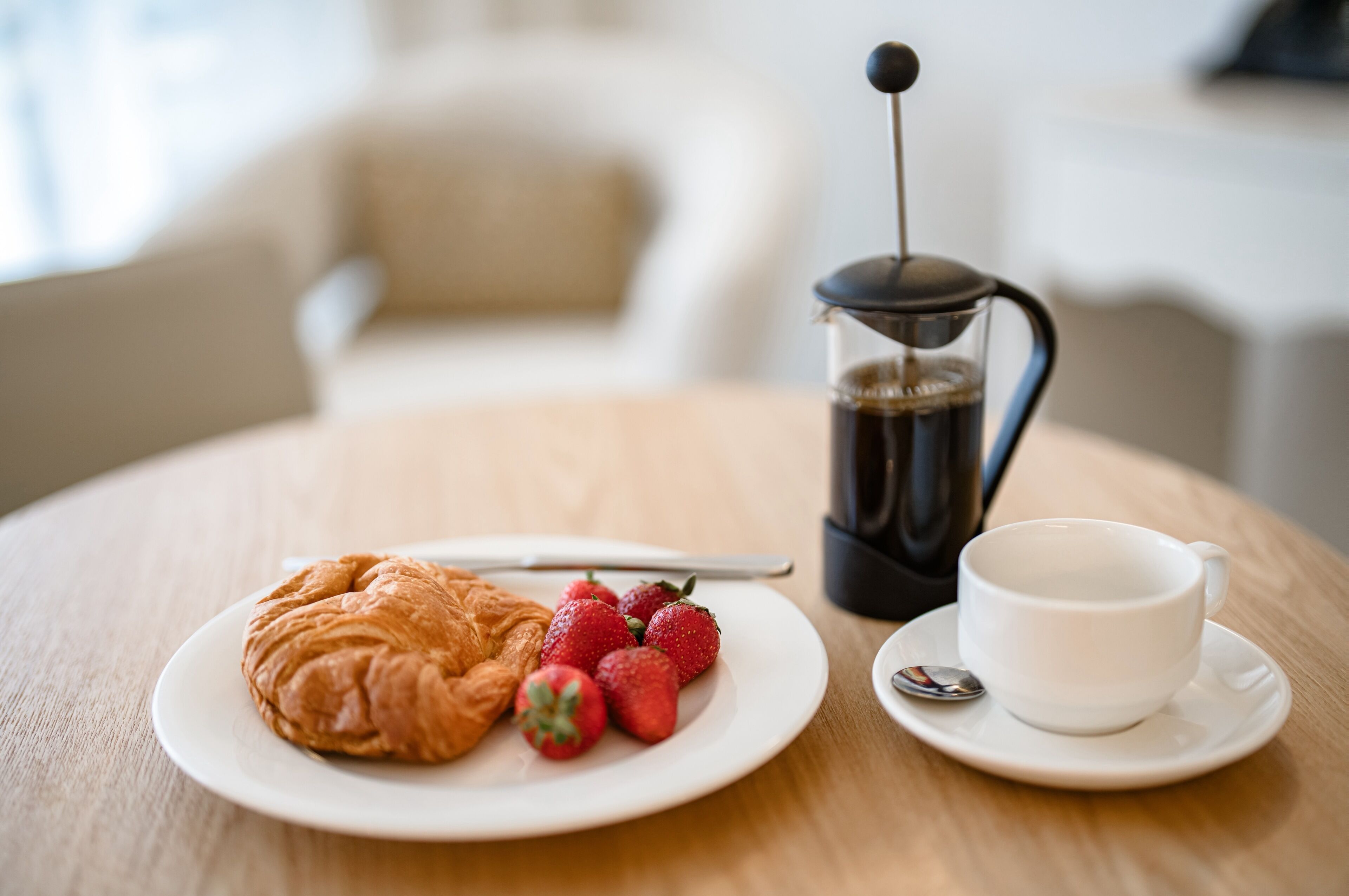 Una escena de desayuno con un croissant, fresas, una cafetera y una taza sobre una mesa de madera.