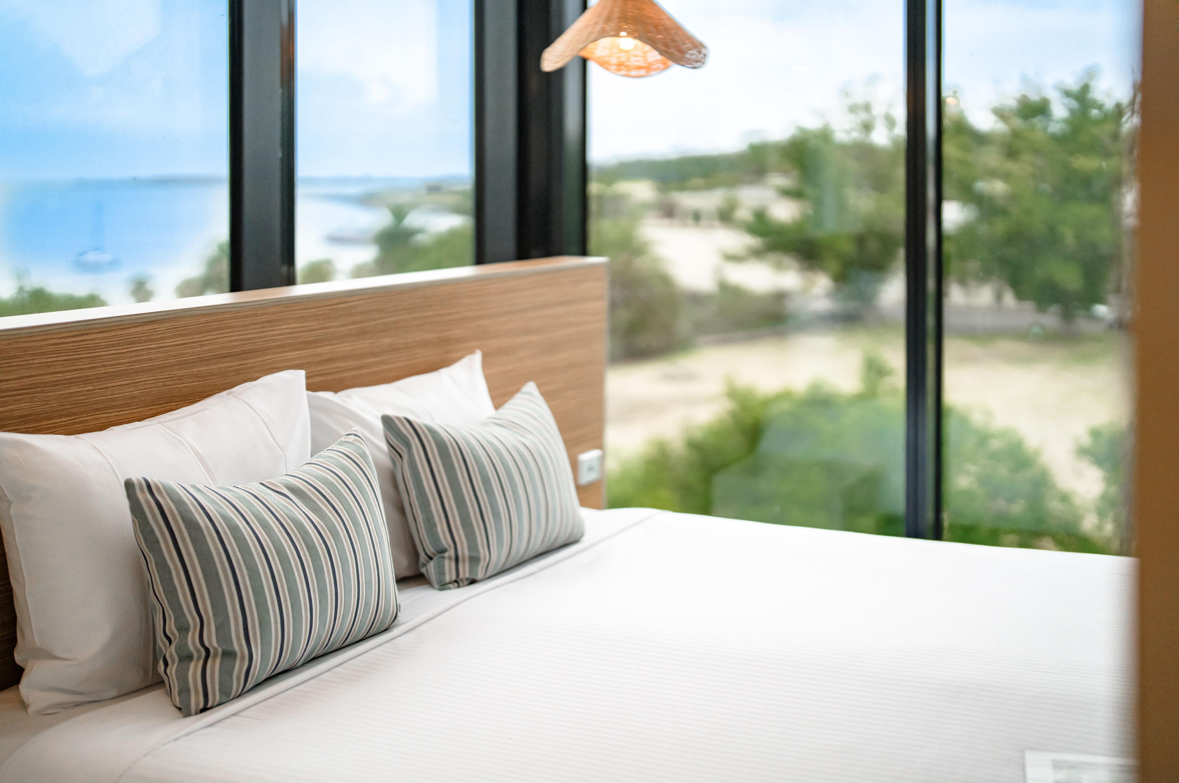 A bed with white bedding and striped pillows, a wooden headboard, and a view of a beach and greenery through large windows.