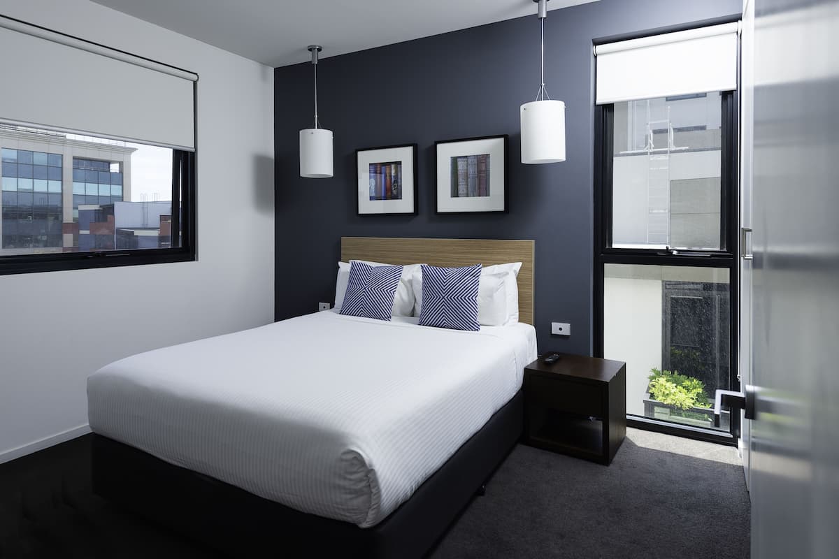 A modern bedroom with a large bed, two framed pictures on the wall, a nightstand, and a window with a view of buildings.