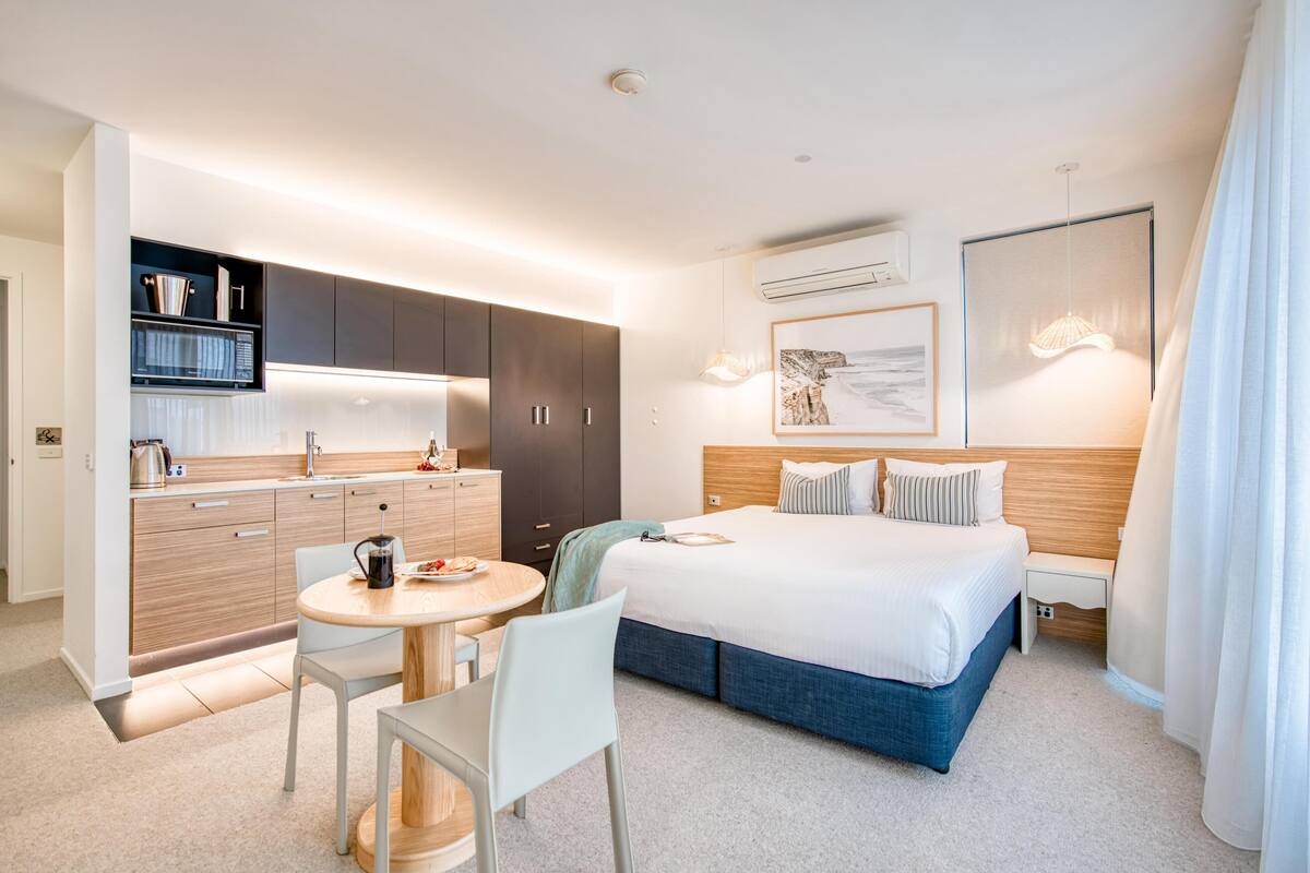 A modern hotel room with a bed, bedside table, and a small kitchen area.