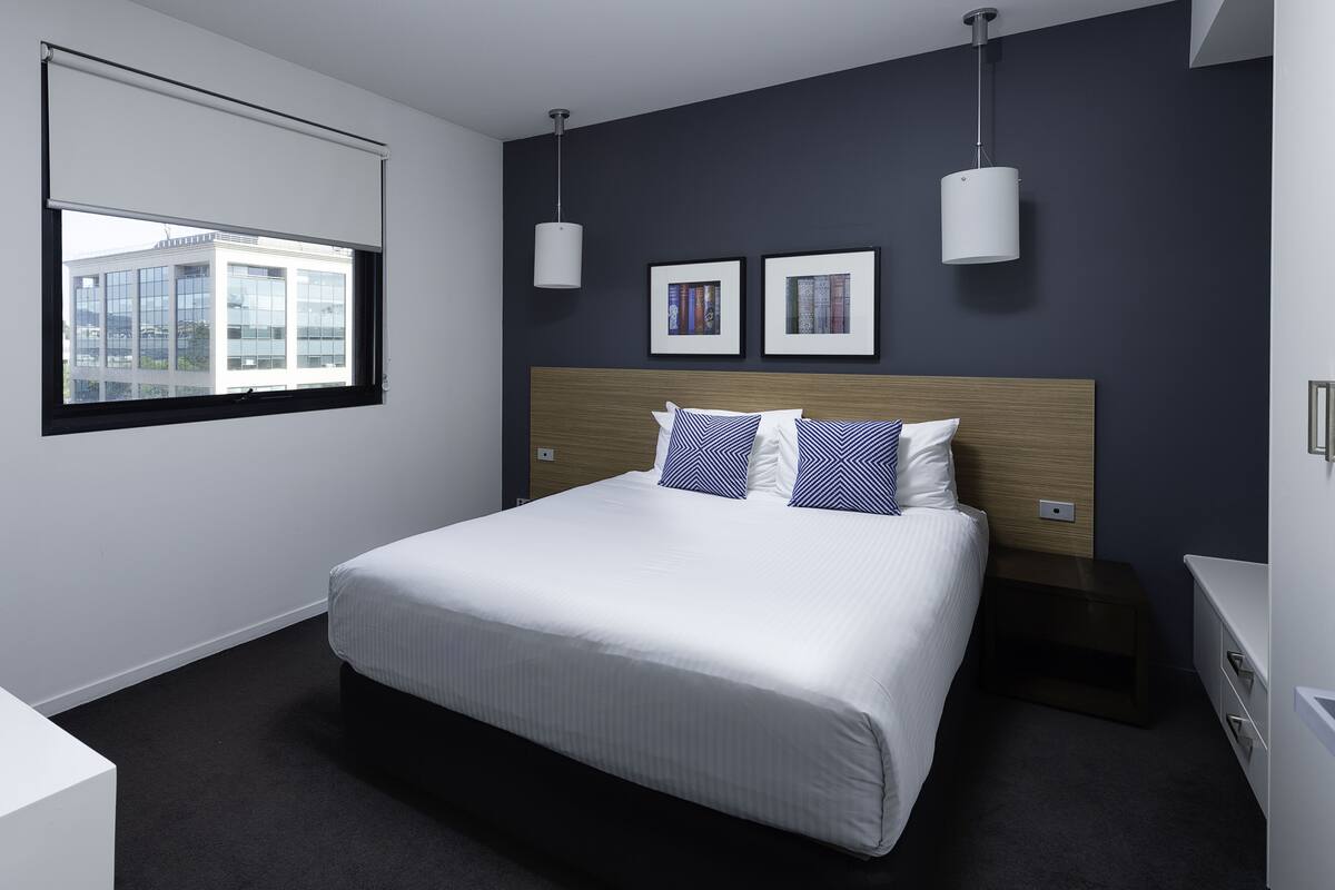 A modern hotel room with a large bed, two bedside tables, a window with blinds, and two wall-mounted lights.