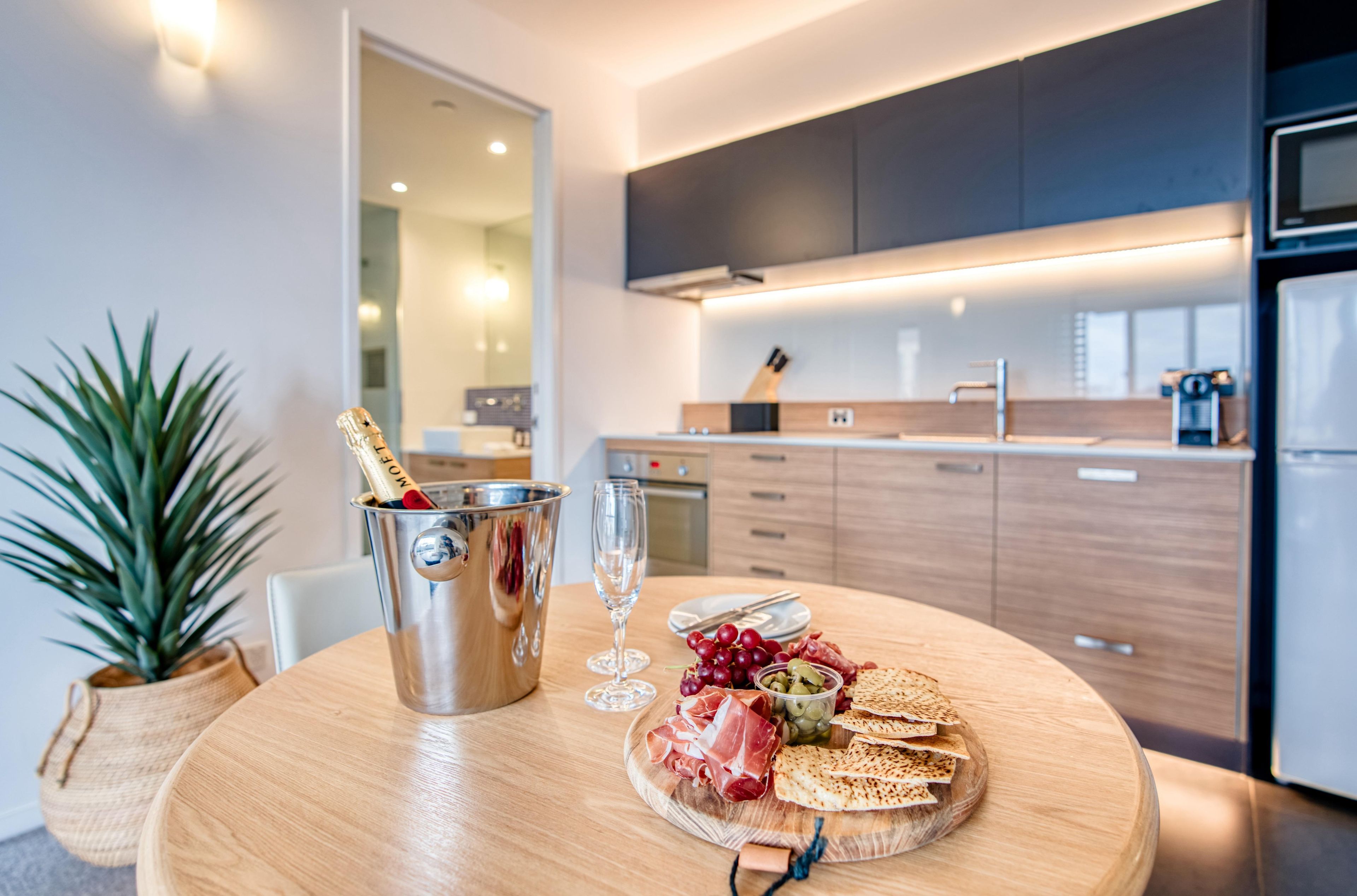 A modern kitchen with a wooden table set for a meal, including a bottle of champagne, a glass, and a plate with food.