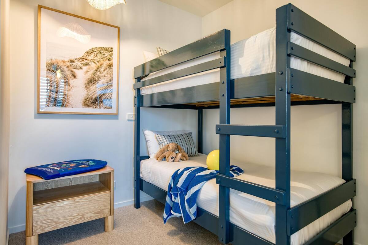 A bunk bed with a teddy bear and a ball, a small wooden shelf, and a framed picture on the wall.