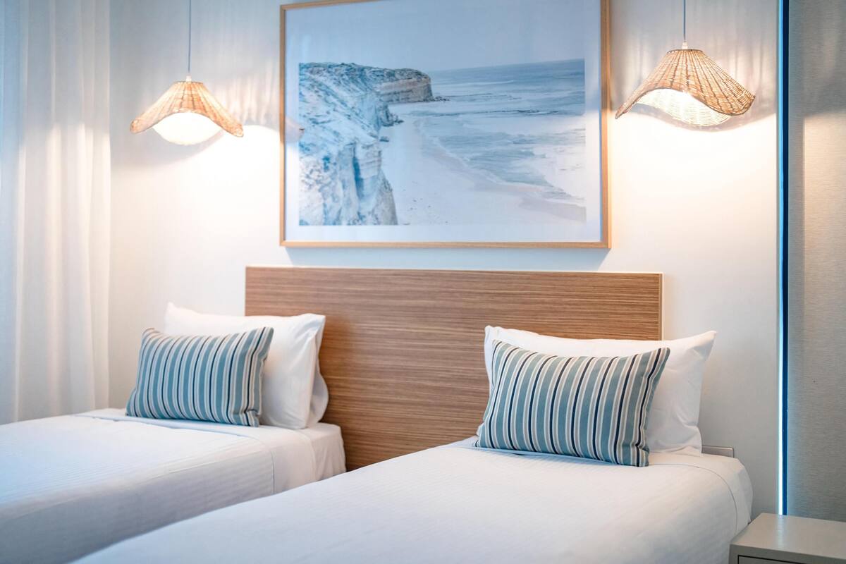 A hotel room with two beds, a wooden headboard, a framed picture of a beach, and two hanging lamps.