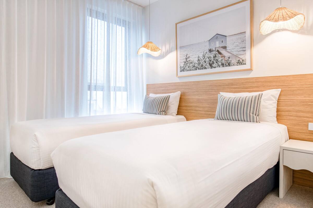 A hotel room with two beds, a large window with sheer curtains, a framed picture on the wall, and two wall-mounted lamps.