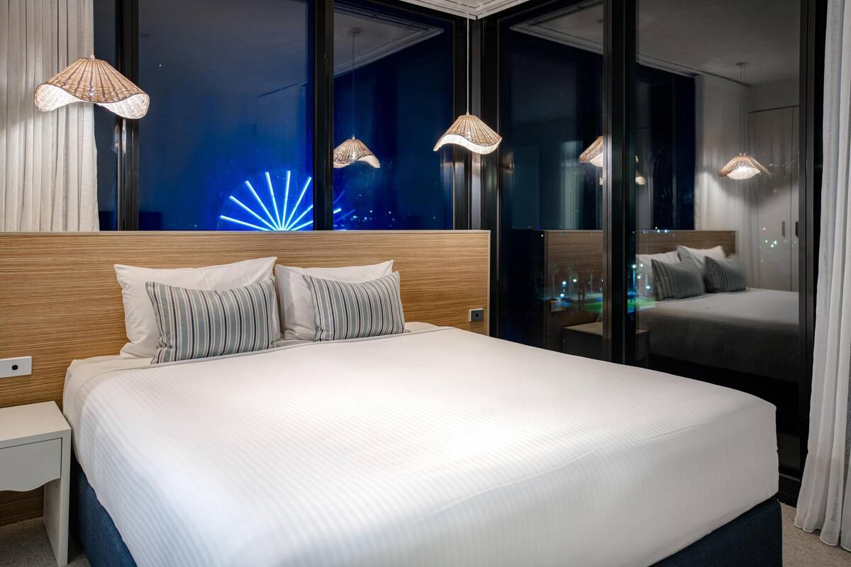 A modern hotel room with a large bed, two bedside tables, and a nightstand. There are three hanging lamps and a view of the city lights through the window.