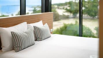 A bed with white bedding and striped pillows, a wooden headboard, and a view of a beach and greenery through large windows.