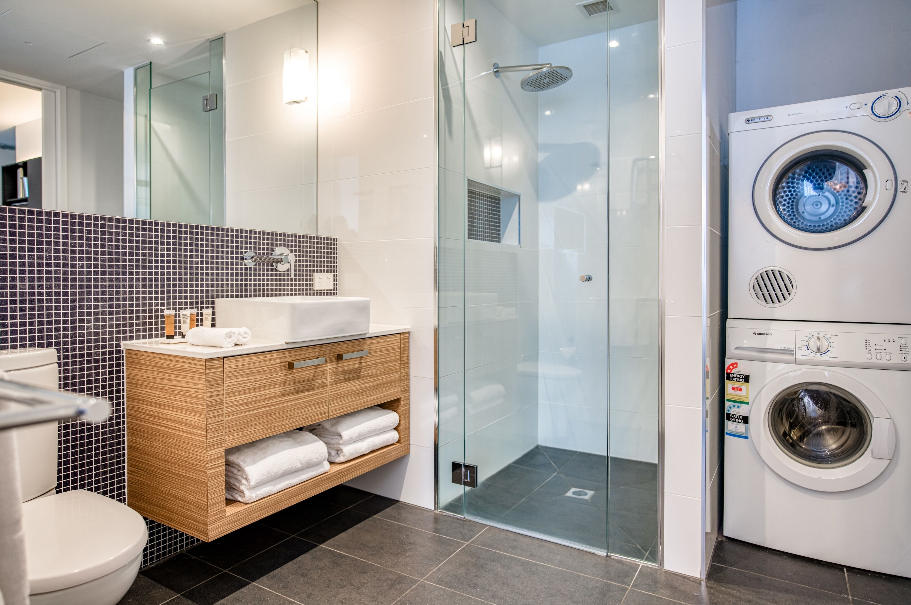 Un baño moderno con cabina de ducha de vidrio, lavadora integrada y tocador de madera con toallas.