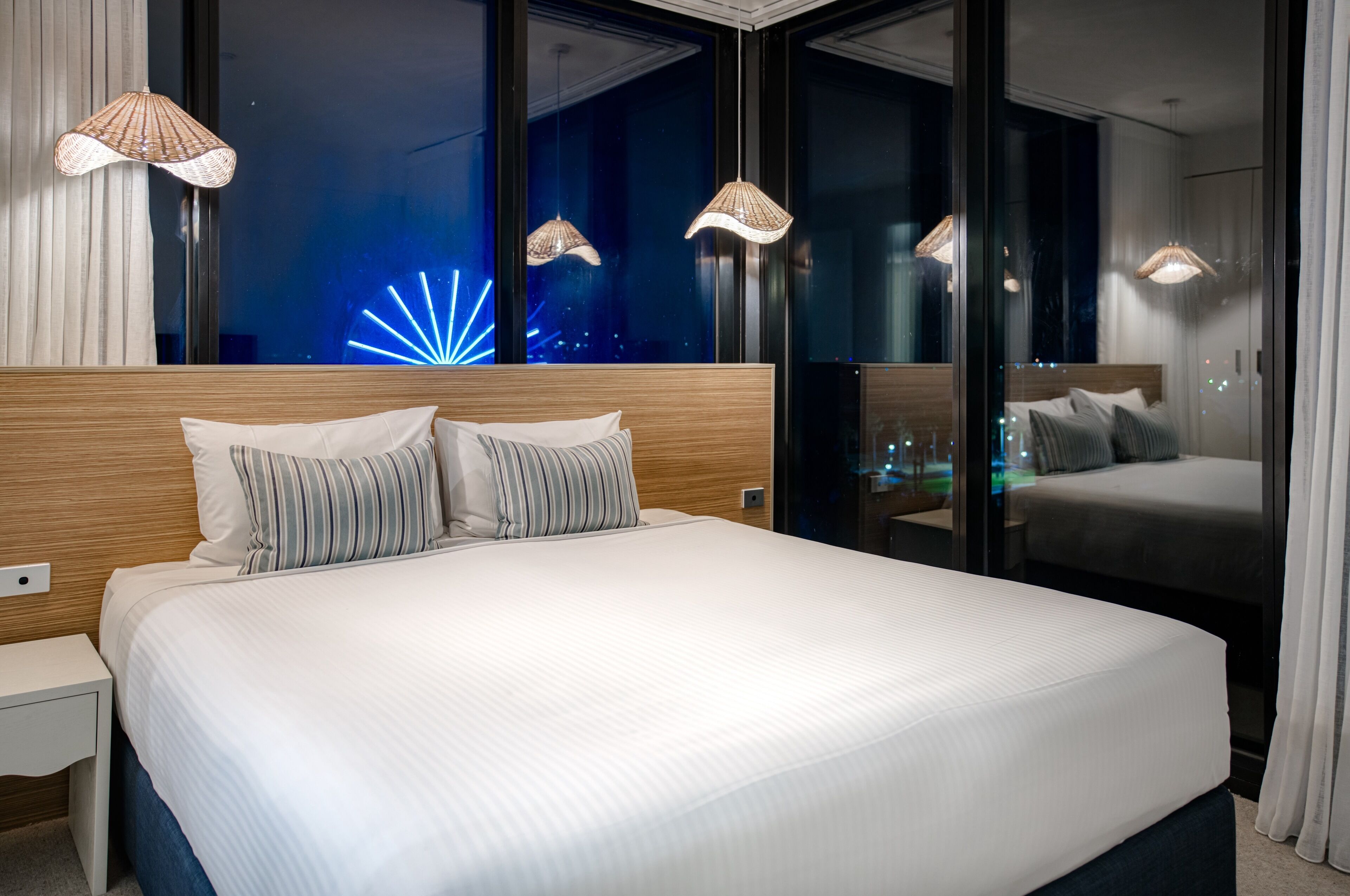 A modern hotel room with a large bed, two bedside tables, and a nightstand. The room features large windows with a view of the city at night, including a visible light display.