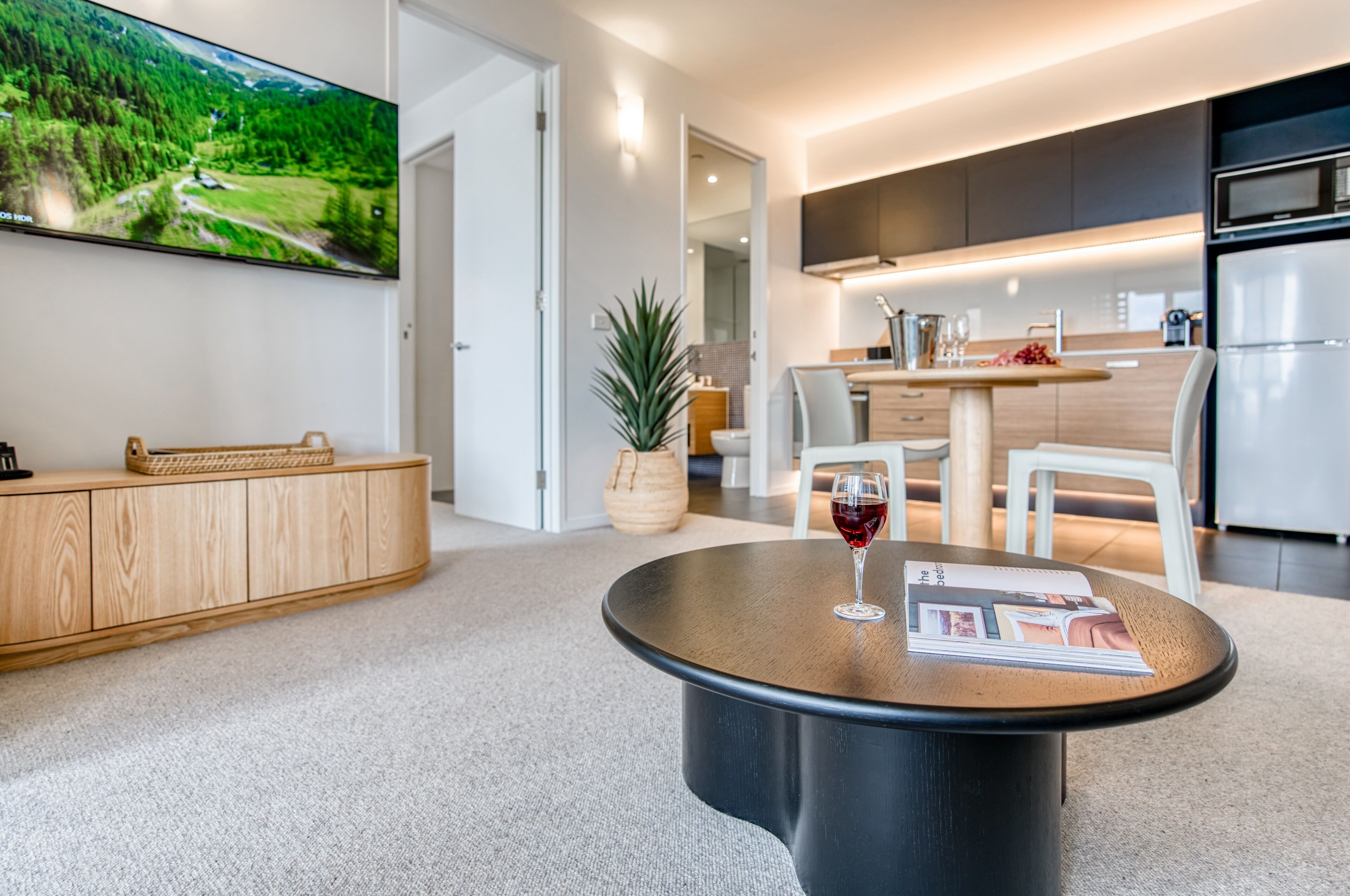 A modern kitchen with a dining area, a large flat-screen TV, and a round coffee table with a magazine and a glass of red wine.