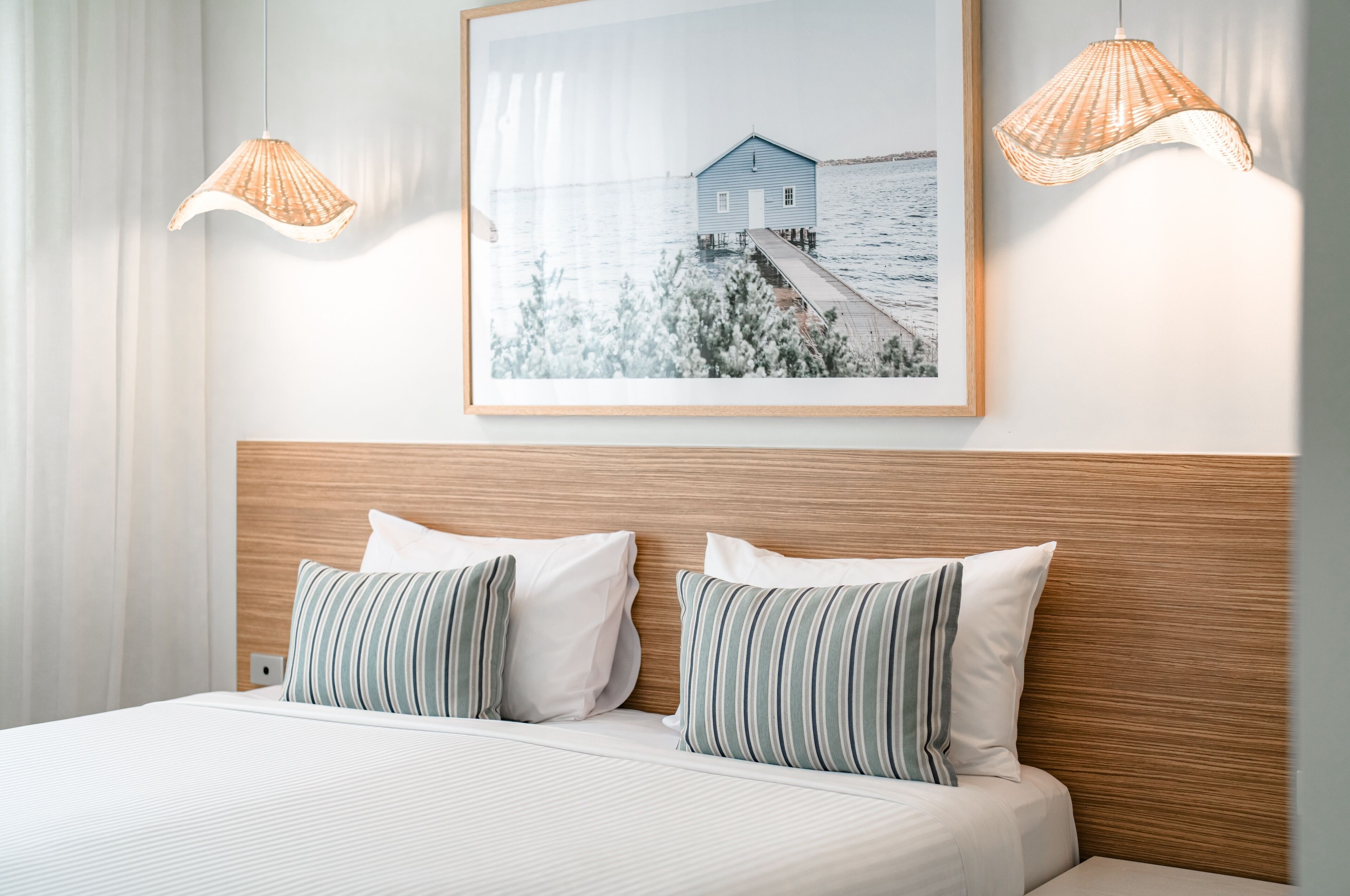 A hotel room with a wooden headboard, two beds with striped pillows, a framed picture of a blue house on water, and two wicker lamps.