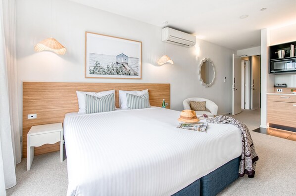 A neatly made bed with a headboard, a bedside table, a wall-mounted air conditioner, a framed picture, and a doorway leading to a bathroom. - Oaks Vue Suites Geelong (Geelong)