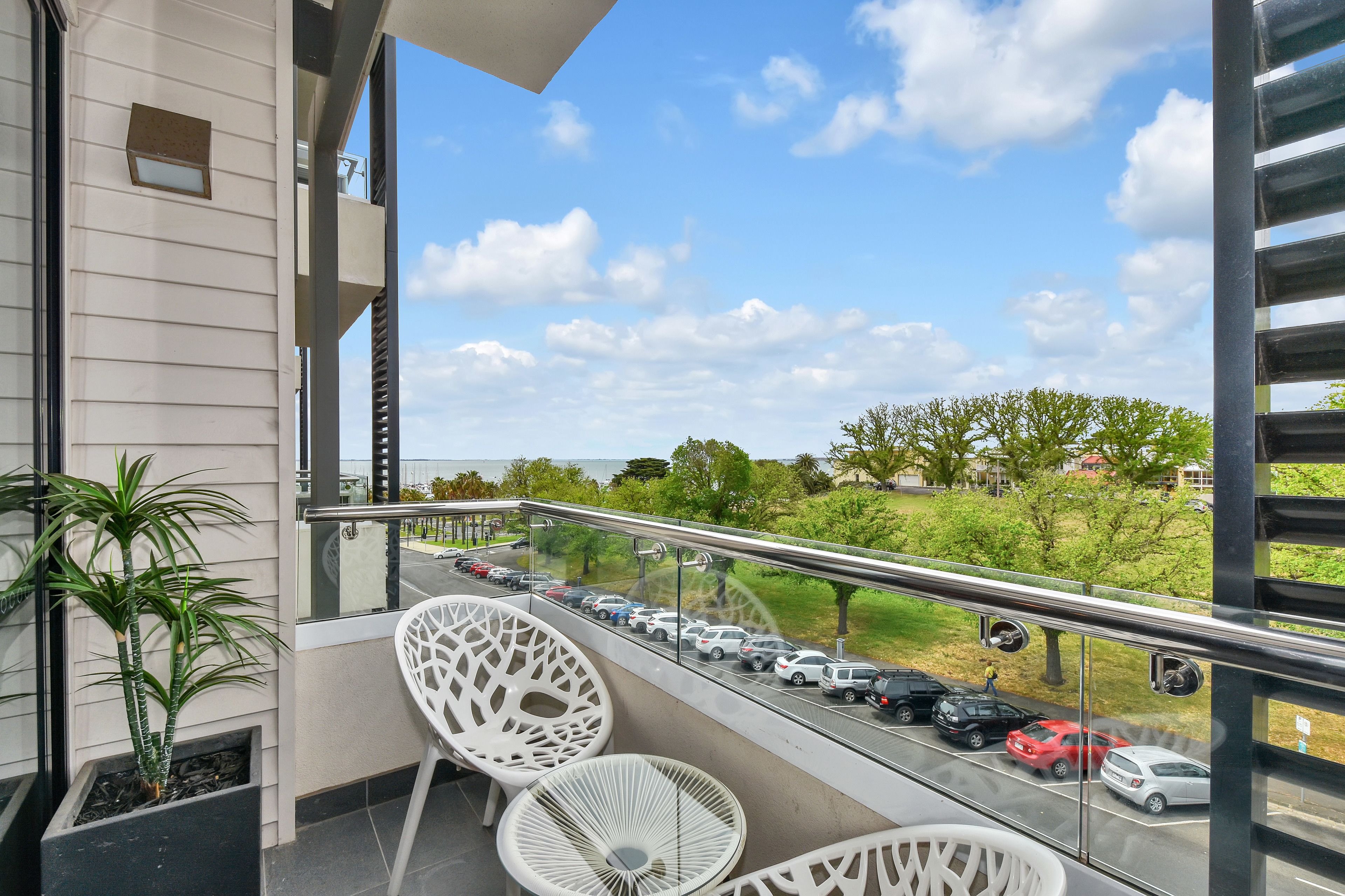 A balcony with two white chairs, a potted plant, and a view of a busy street with cars and greenery.