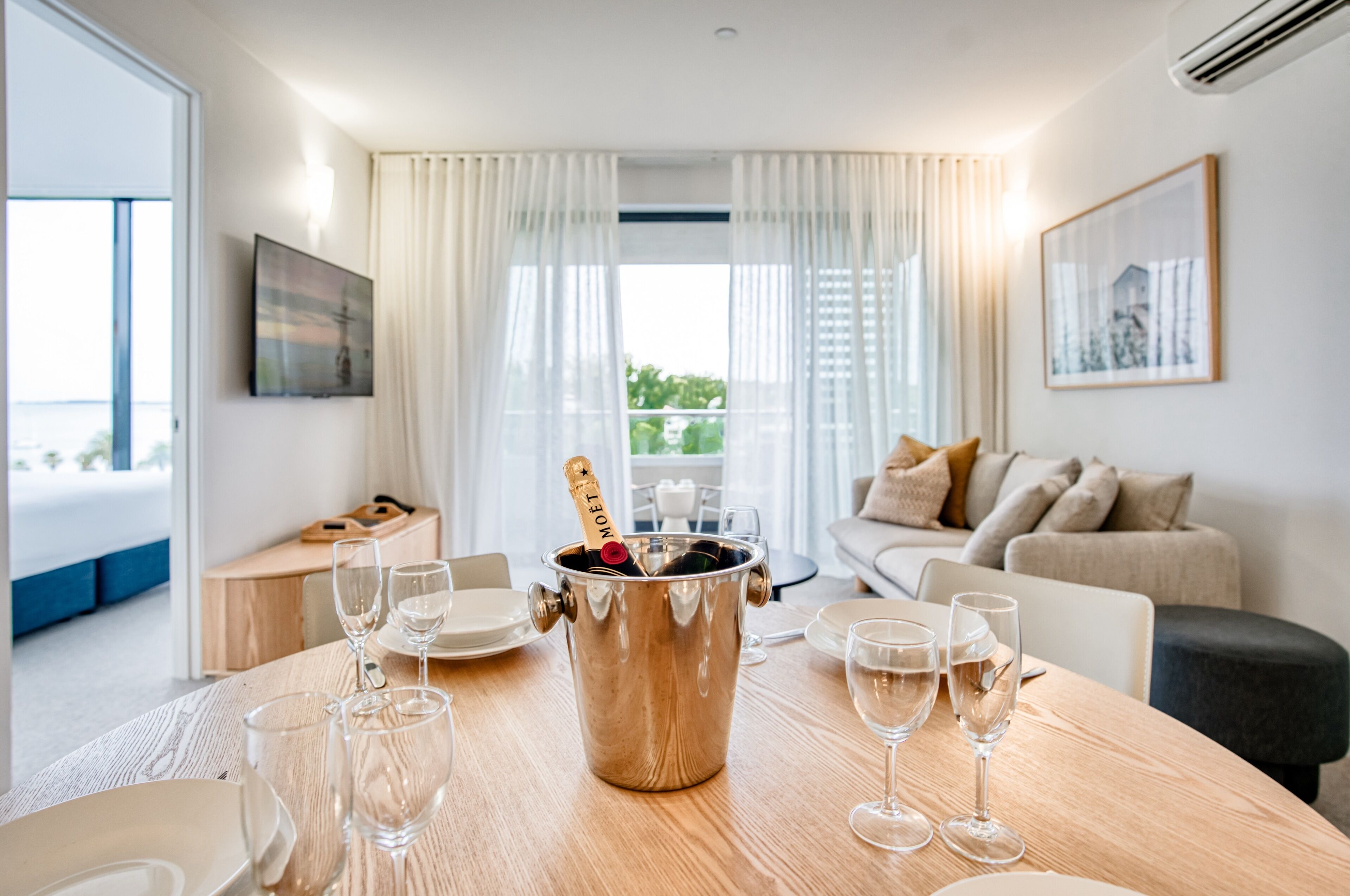 A modern living room with a wooden table set for a celebration, a champagne bucket, and a view of the outdoors through large windows.