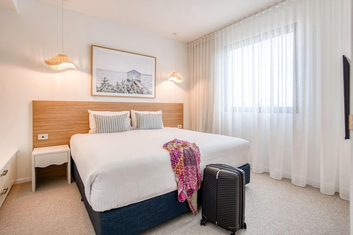 A hotel room with a large bed, a nightstand, a lamp, a picture on the wall, and a window with curtains.