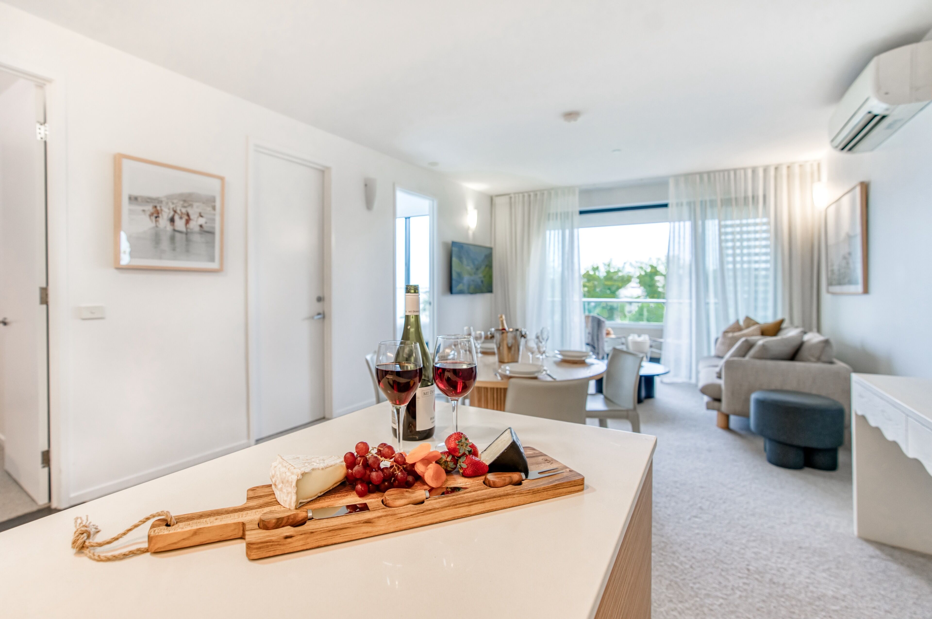 A modern living room with a dining area, featuring a wooden table with a cheese board, wine glasses, and a bottle.