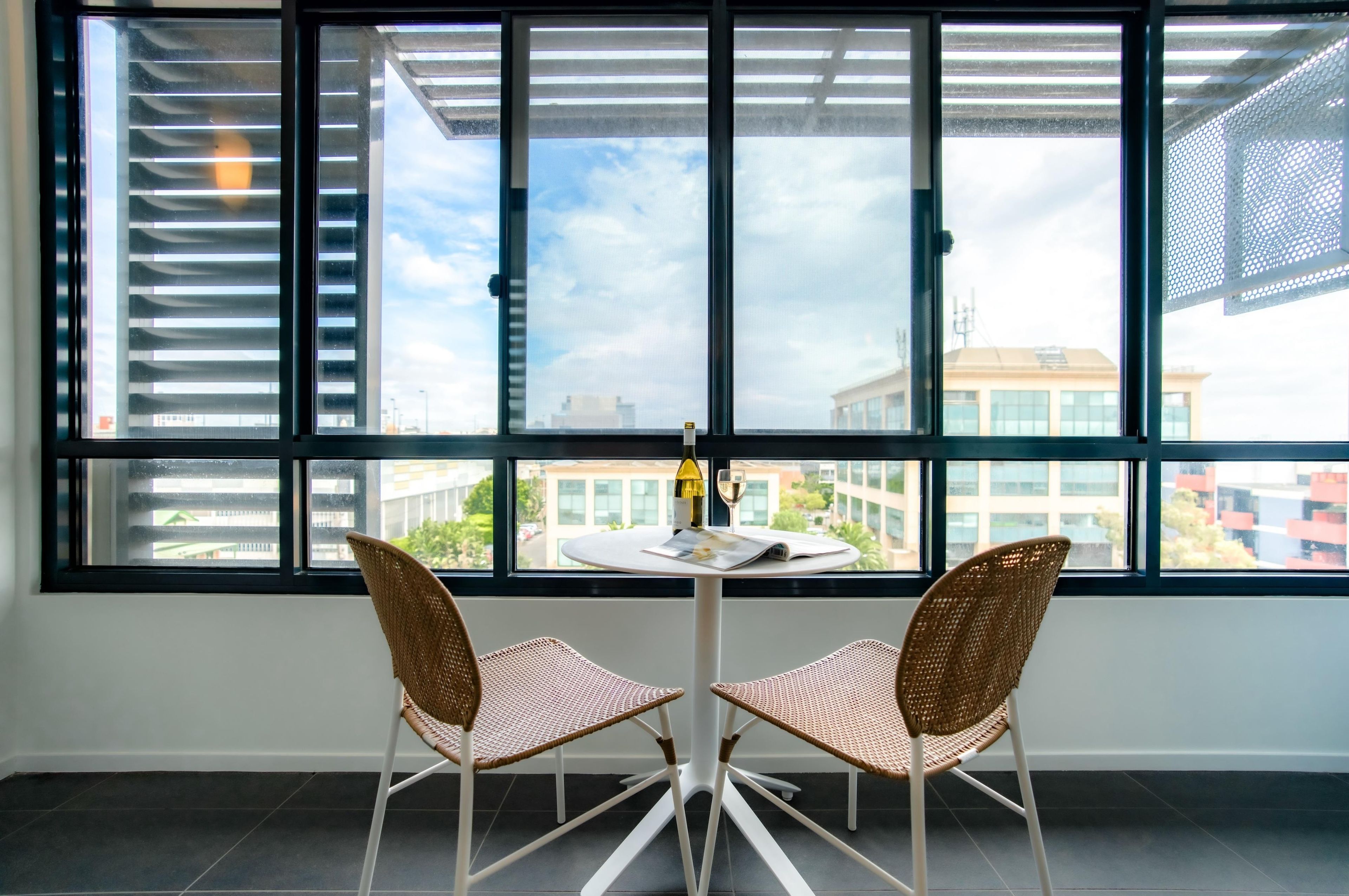 A room with a large window, two wicker chairs, a small table, and a bottle of wine.
