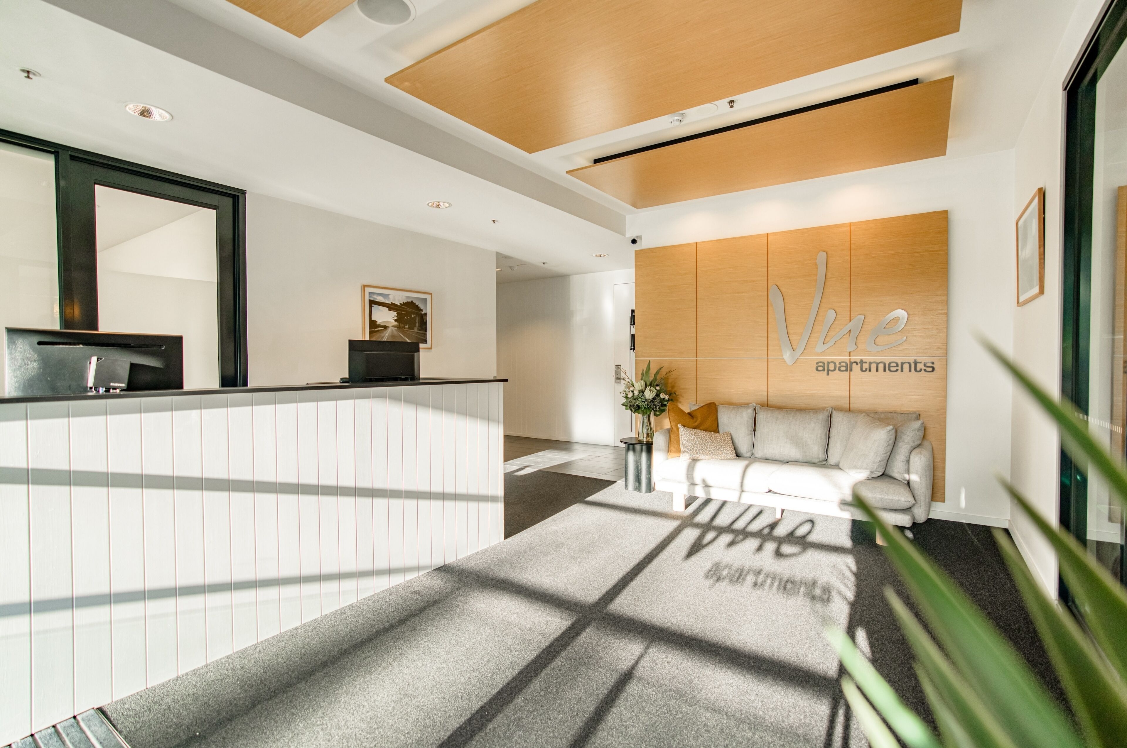 Un espace d’accueil moderne comprenant un comptoir de réception, un canapé et un panneau mural indiquant les appartements Vue.