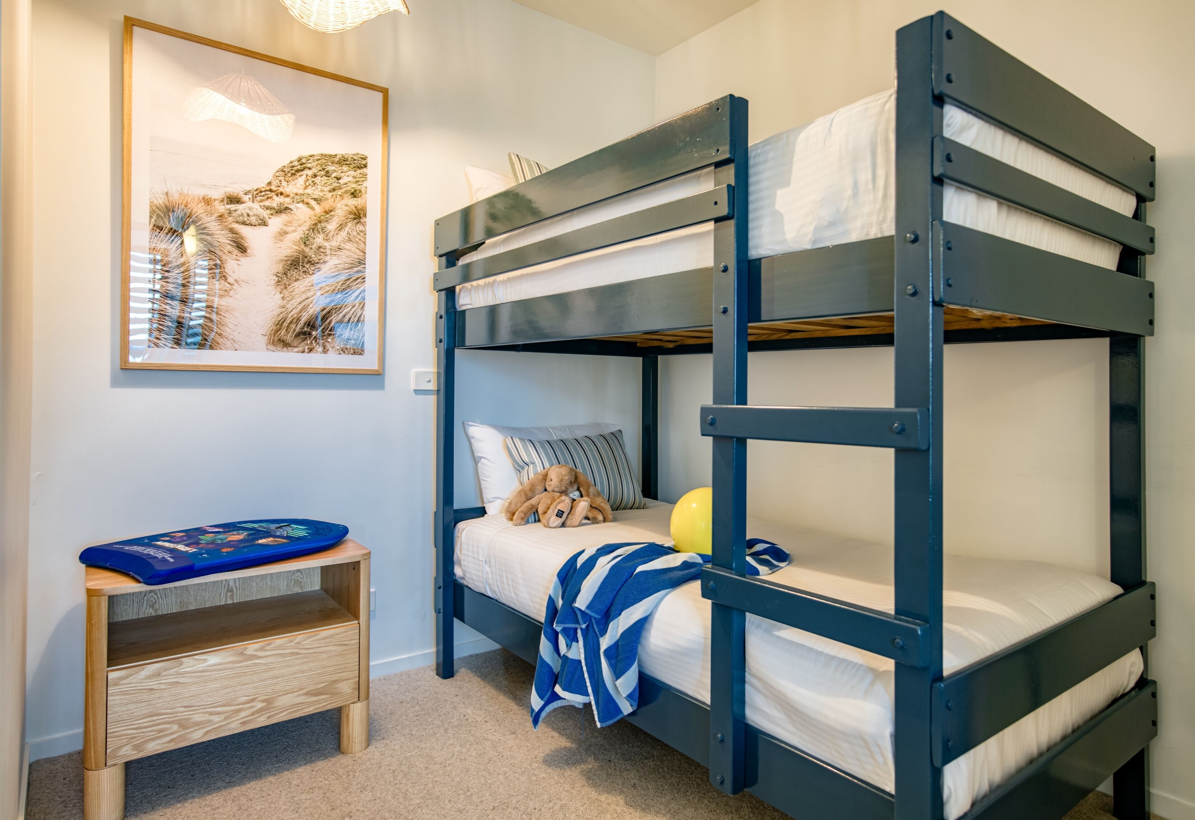 A bunk bed with a teddy bear and a ball, a small wooden table, and a framed picture on the wall.