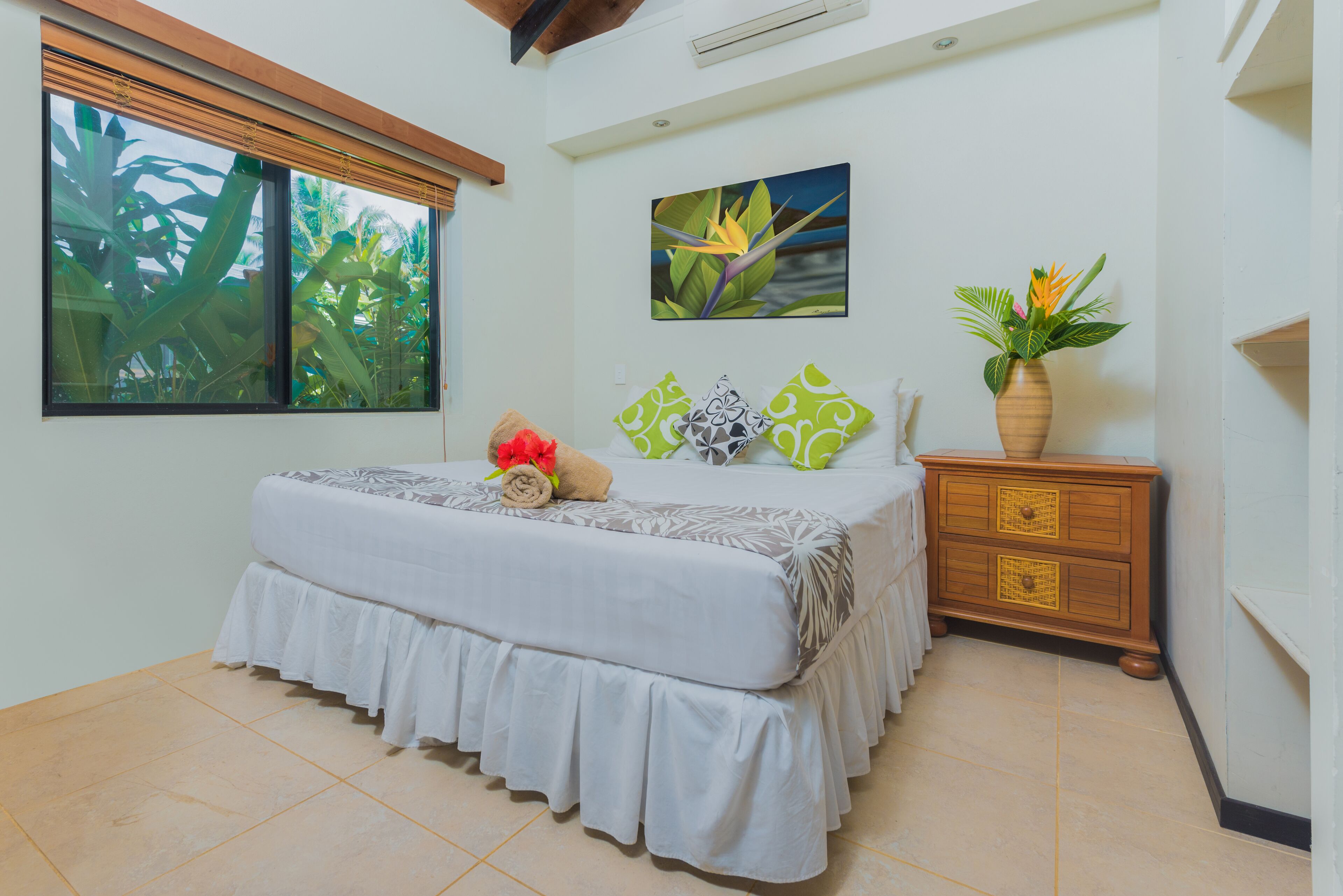 A bedroom with a bed, a window with blinds, a painting, a dresser, and a potted plant.