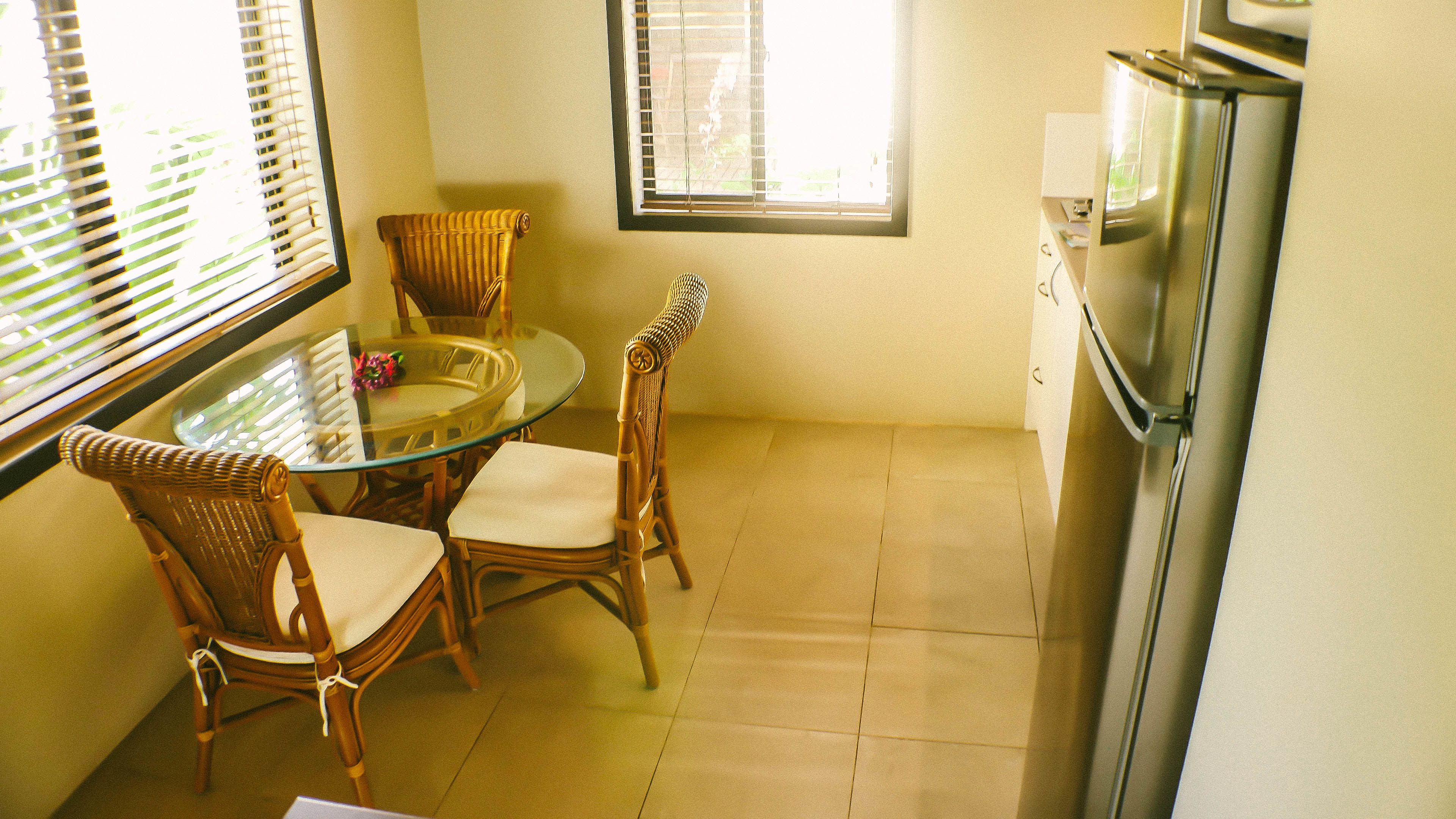 Cocina compacta con una mesa de vidrio, sillas de mimbre, un refrigerador y una ventana con persianas.