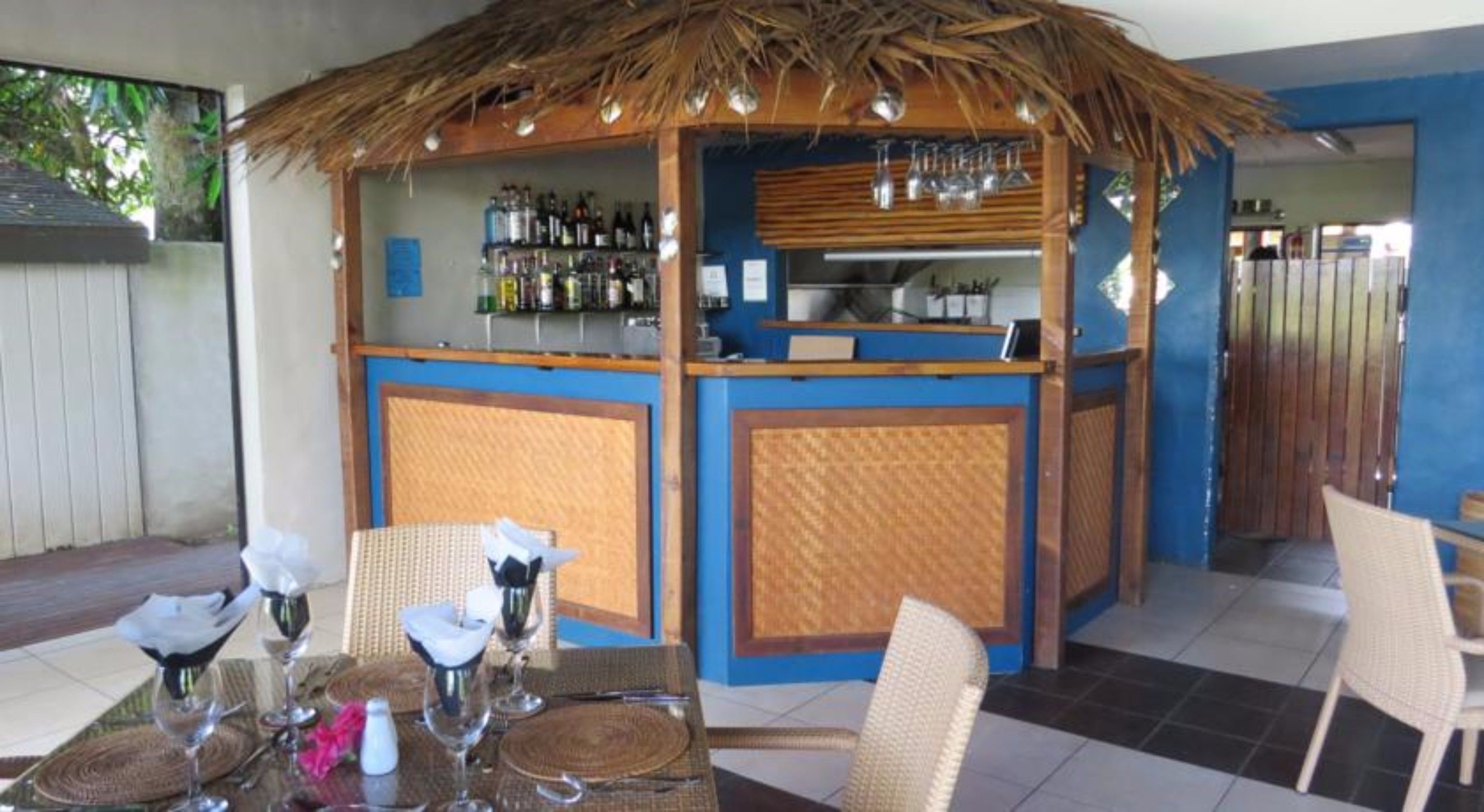 Un área de bar al aire libre con techo de paja, una barra repleta de botellas y mobiliario de mimbre.