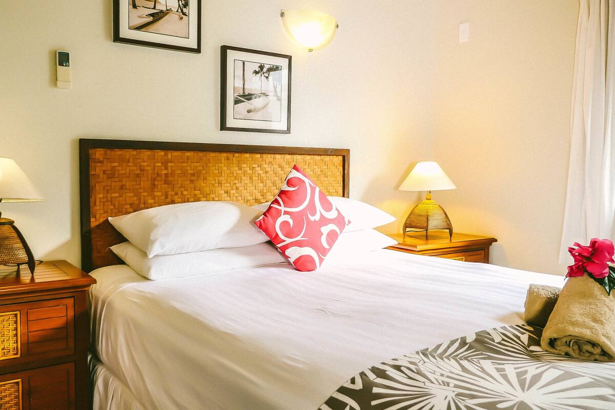 Una cama bien tendida con sábanas blancas, un cojín rojo y blanco, y una manta estampada. Hay dos lámparas de noche y una lámpara empotrada en la pared.