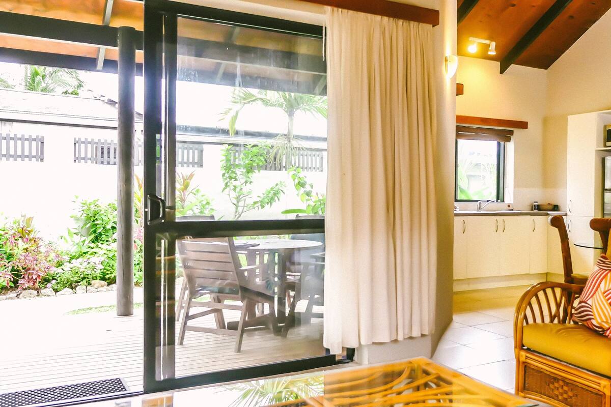 Una habitación con puerta corrediza de vidrio que da a un patio, una silla de mimbre con cojines, una mesita y un microondas en la zona de la cocina.