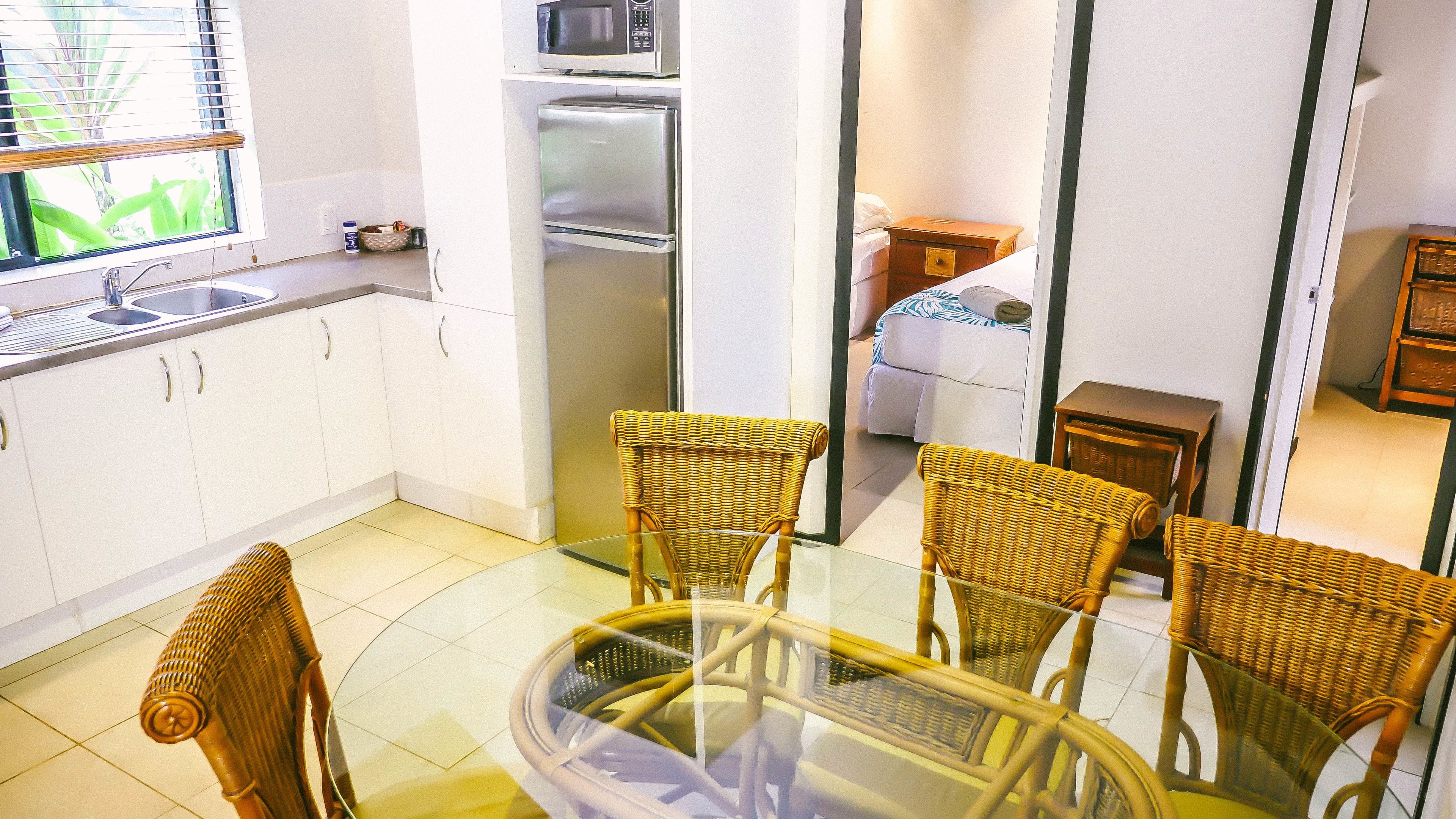 A compact kitchen with white cabinets, a stainless steel refrigerator, a microwave, and a glass dining table with wicker chairs.