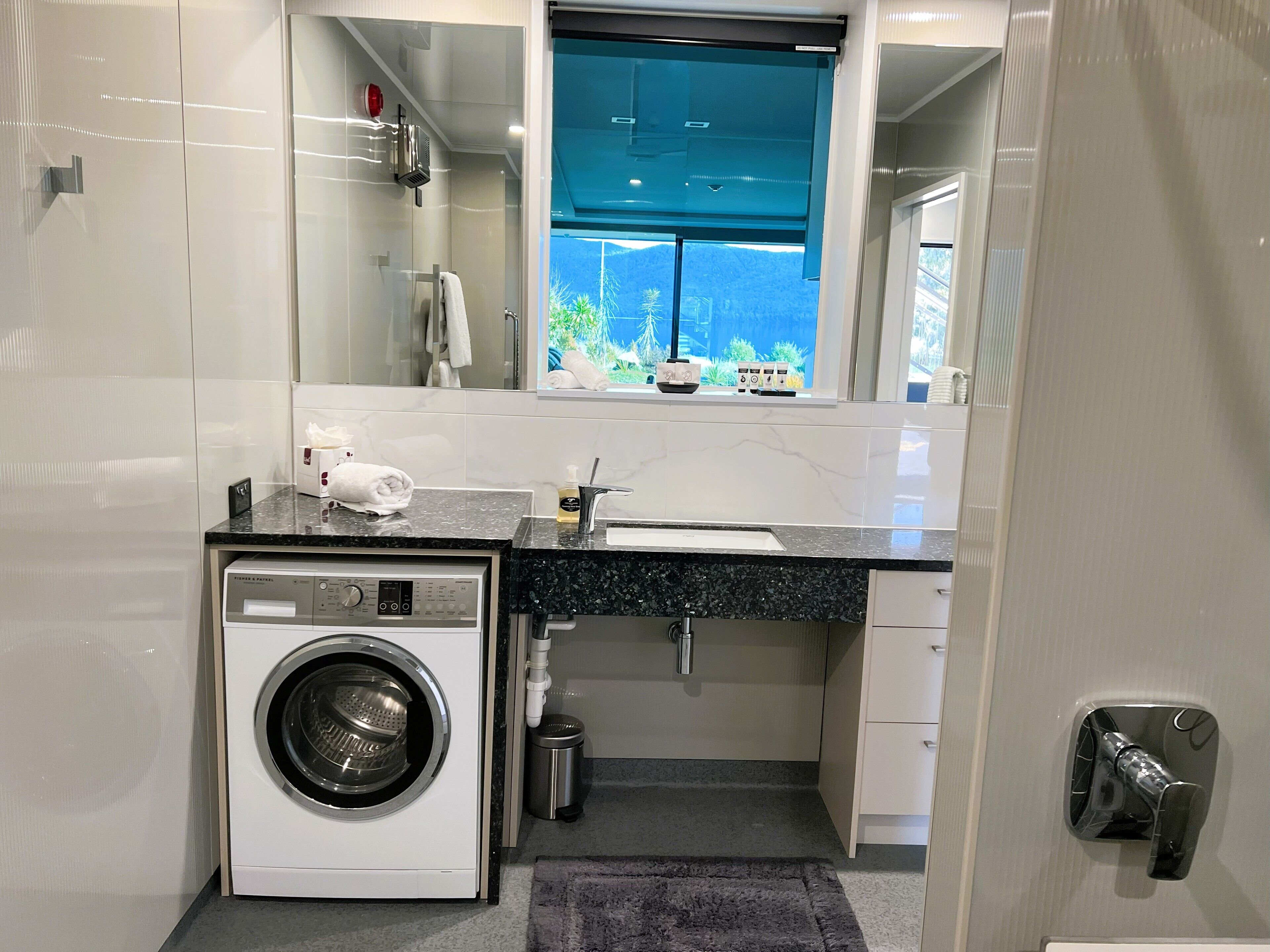 Een compacte, moderne badkamer met een ingebouwde wasmachine en droger, een wastafel met spiegel erboven en een raam met uitzicht op groen.