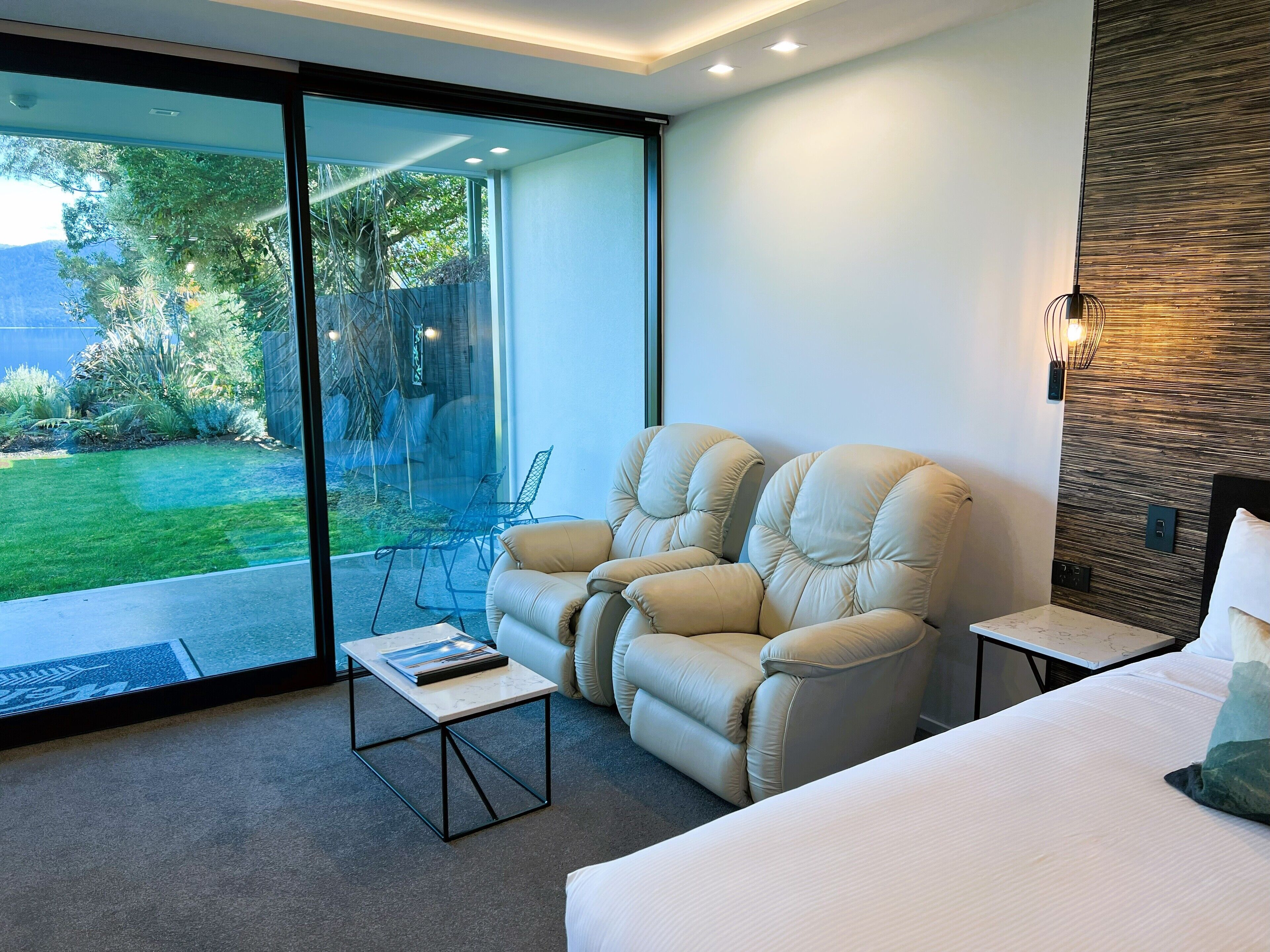 A modern bedroom with a large bed, a reclining chair, a small table, and a view of a garden through a sliding glass door.